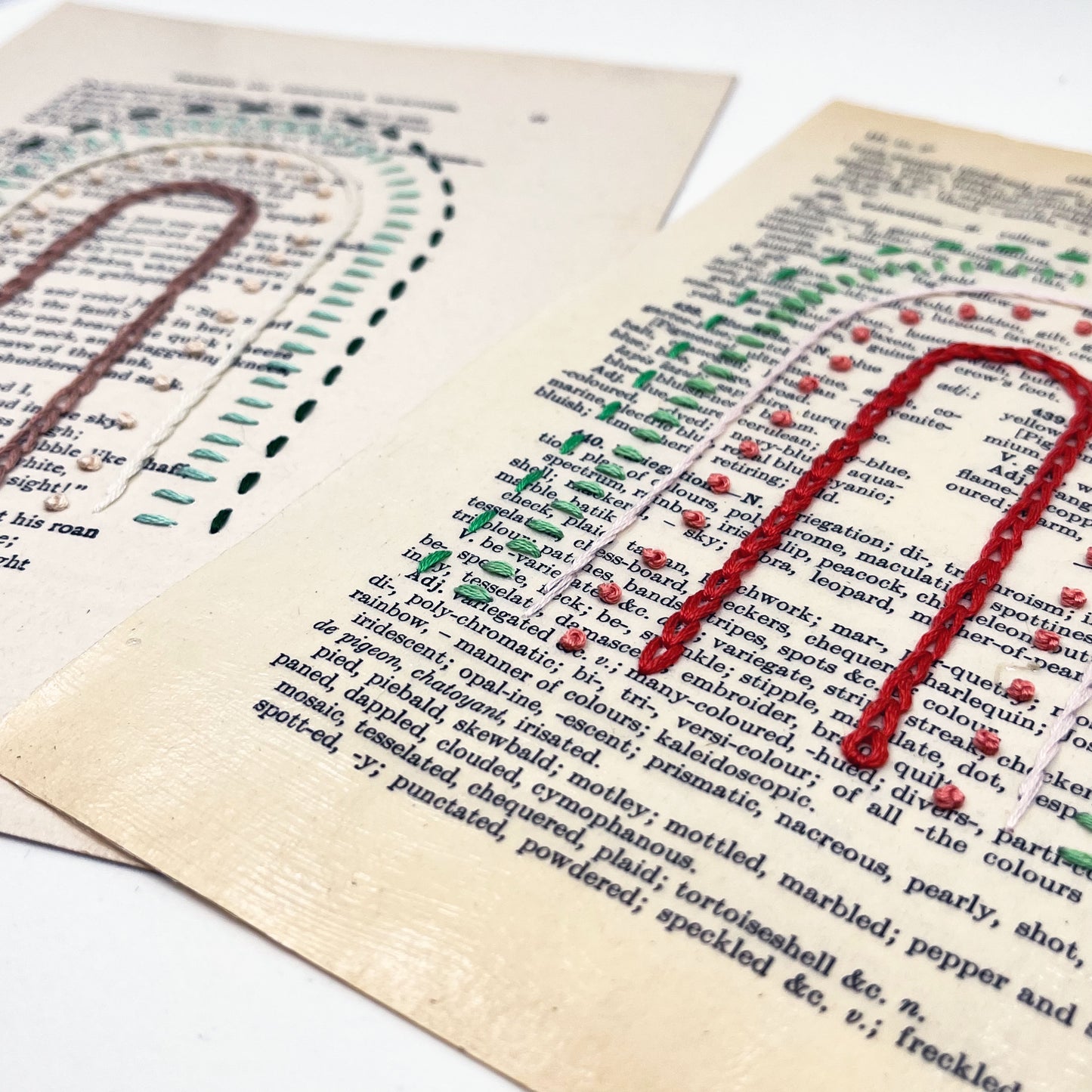 a close up angled view of two vintage book pages, embroidered with a rainbow in different types of embroidery stitches, on a white background