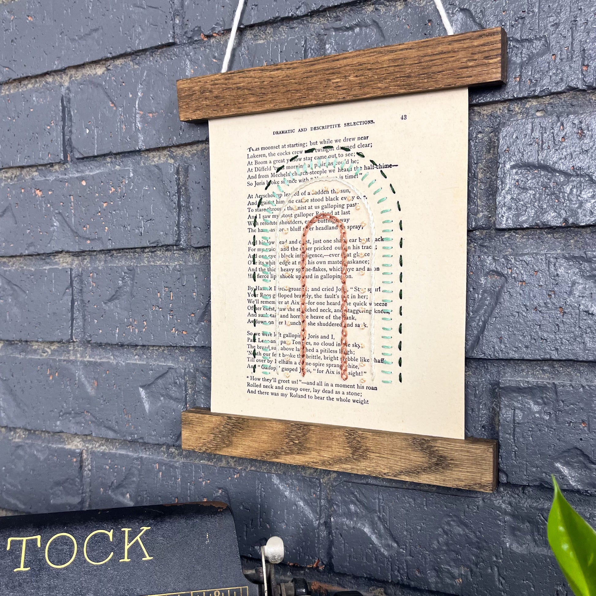 Vintage thesaurus page, embroidered with a rainbow in different types of stitches. Hanging in a magnetic wood frame on a grey brick wall, with the top of a typewriter and pothos plant peeking out at the bottom of the picture