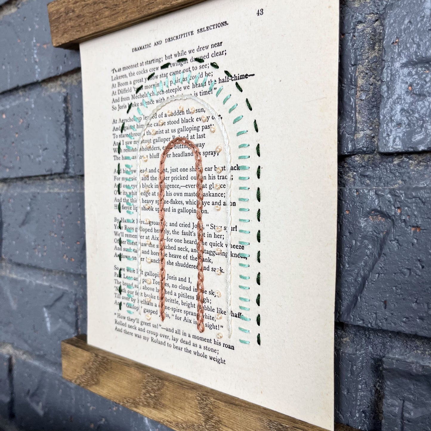 close up angled view of a vintage book page, embroidered with a rainbow of mauve, peach, ivory and greens in different types of stitches. Hanging in a magnetic wood frame on a grey brick wall