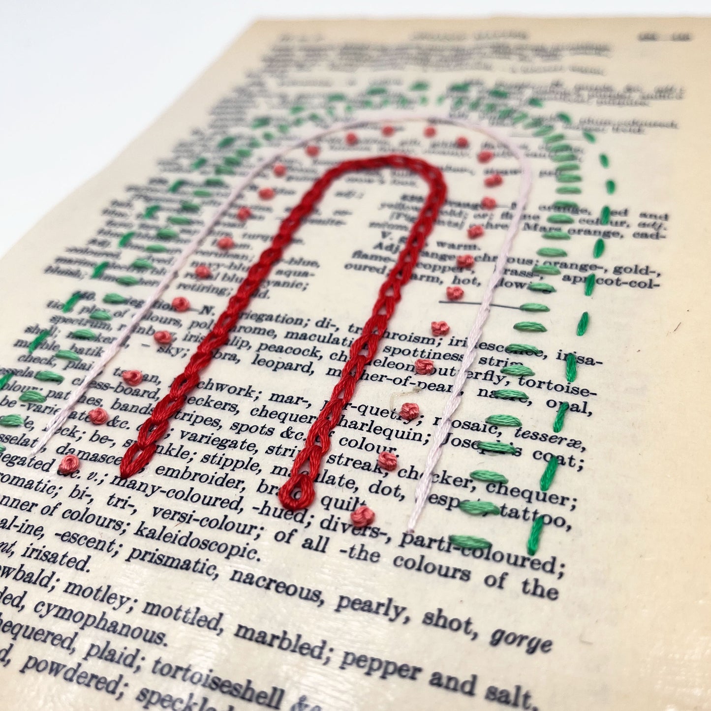 a close up angled view of a vintage book page, embroidered with a rainbow of red, coral, peach and greens in different types of embroidery stitches, on a white background