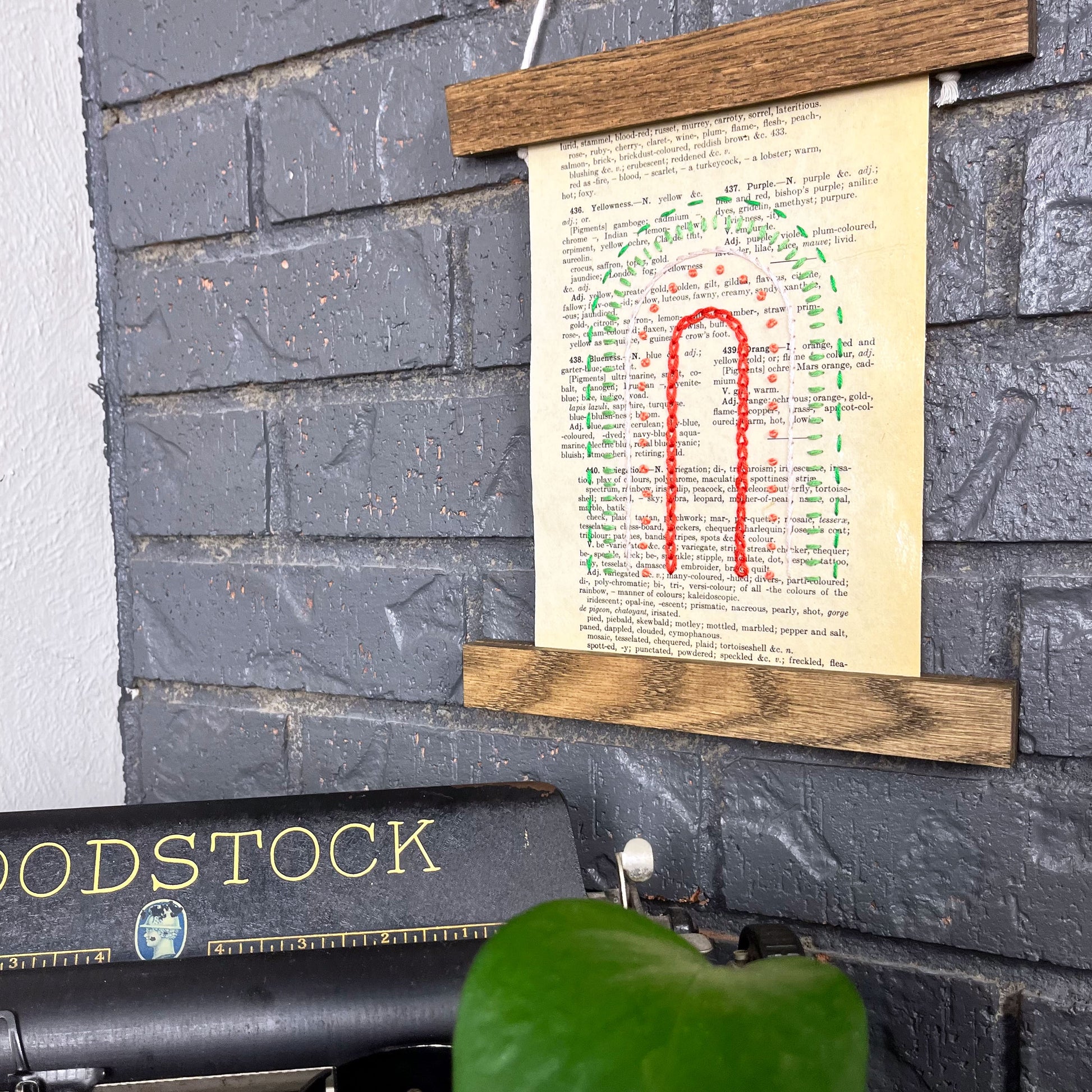 Vintage thesaurus page, embroidered with a rainbow in different types of stitches. Hanging in a magnetic wood frame on a grey brick wall, with the top of a typewriter and pothos plant peeking out at the bottom of the picture
