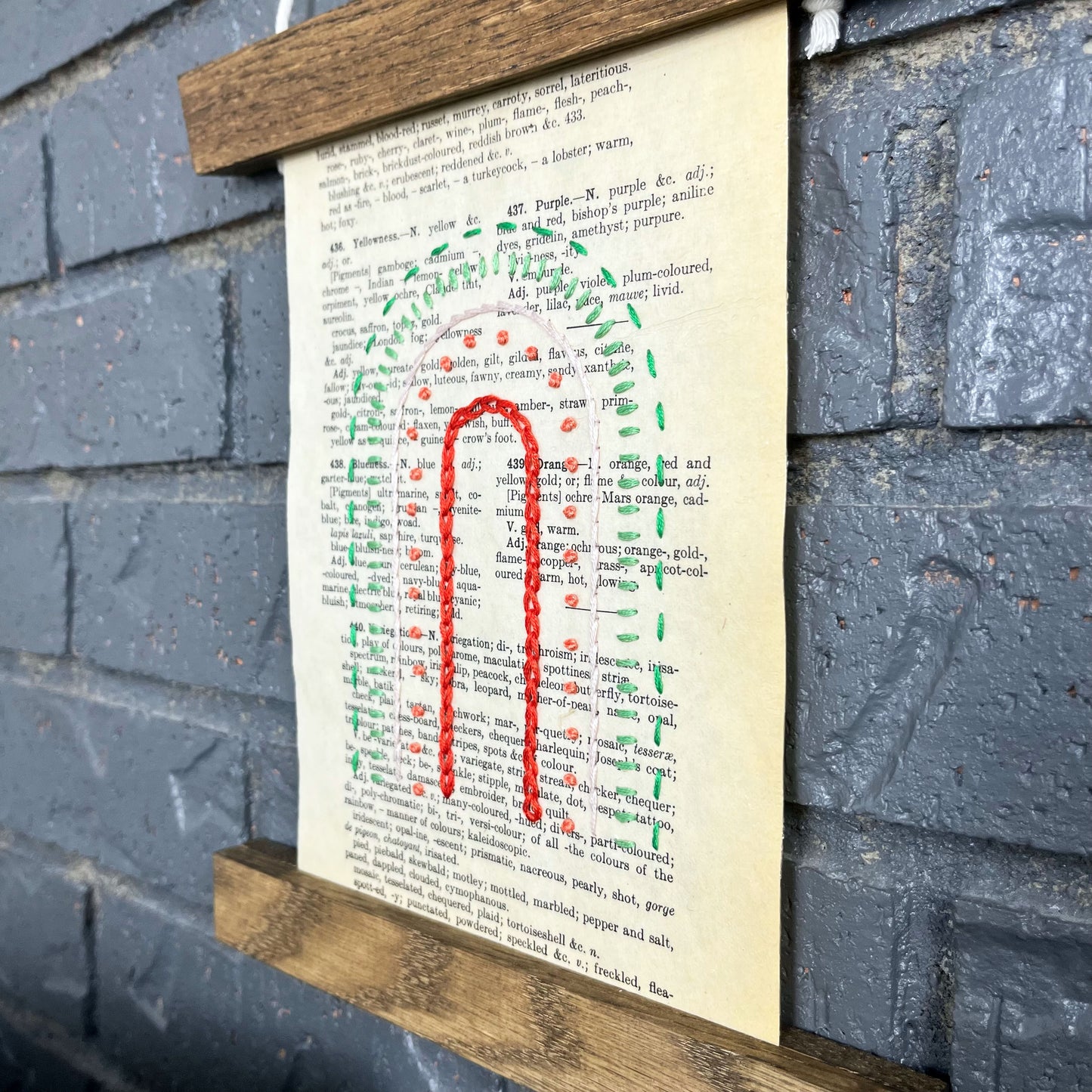 close up angled view of a vintage book page, embroidered with a rainbow of red, coral, peach and greens in different types of stitches. Hanging in a magnetic wood frame on a grey brick wall