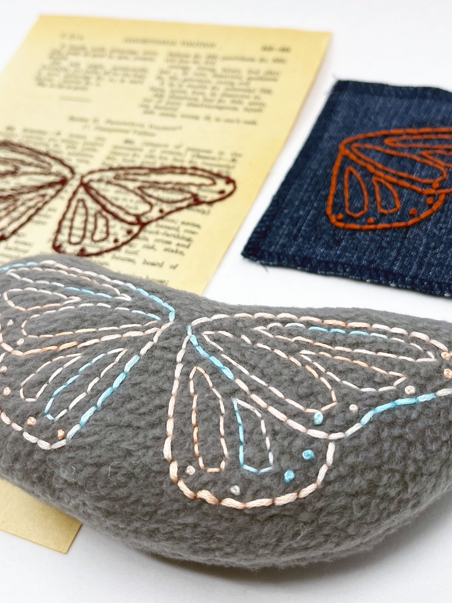 a group of an old book page from a thesaurus, a denim patch, and a grey stuffie with butterfly wings hand embroidered on them