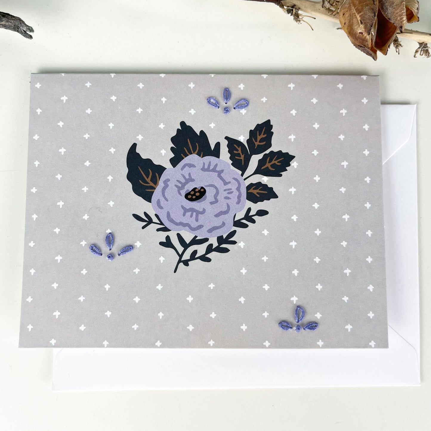 gray and purple floral greeting card embellished with small hand embroidered flowers on a white counter