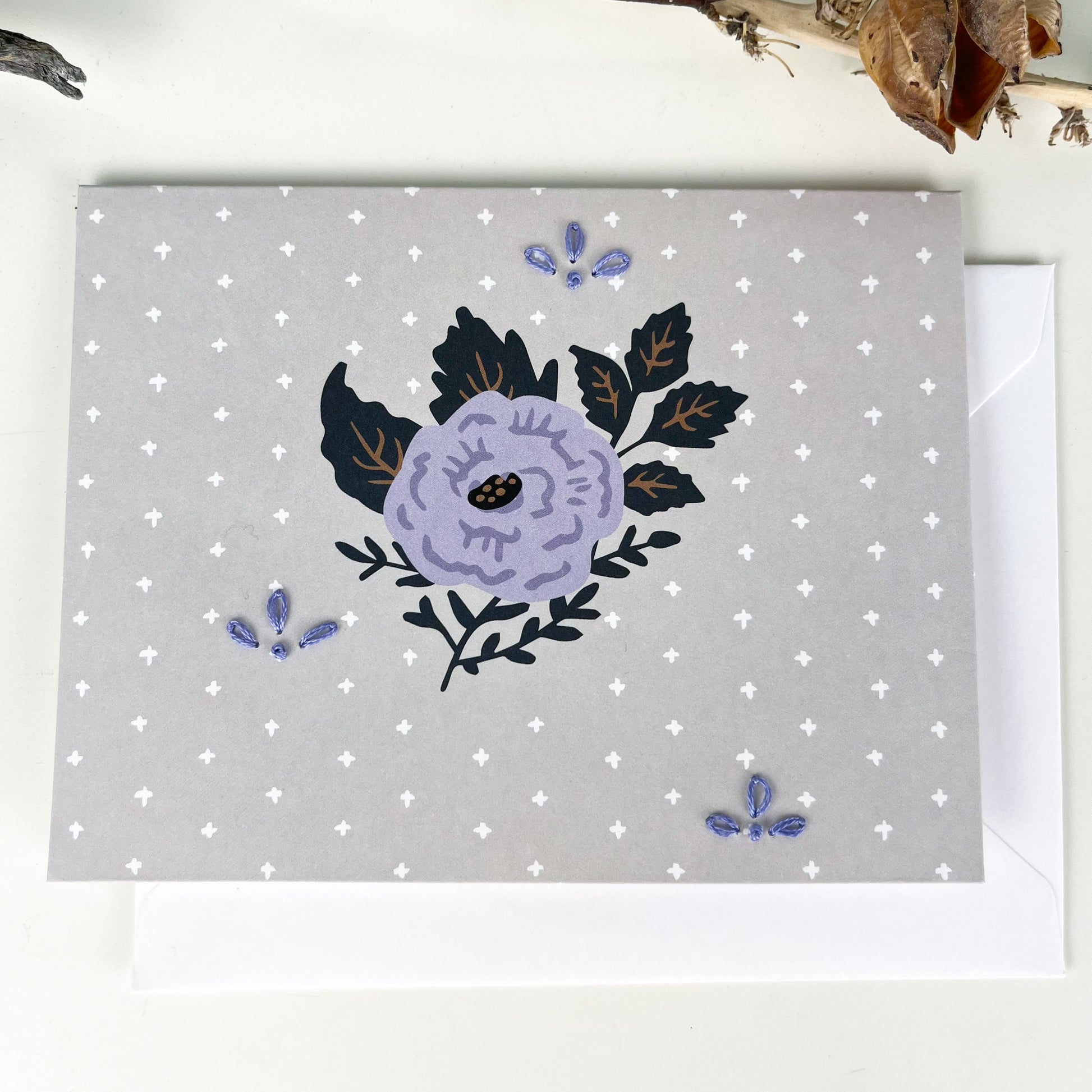 gray and purple floral greeting card embellished with small hand embroidered flowers on a white counter