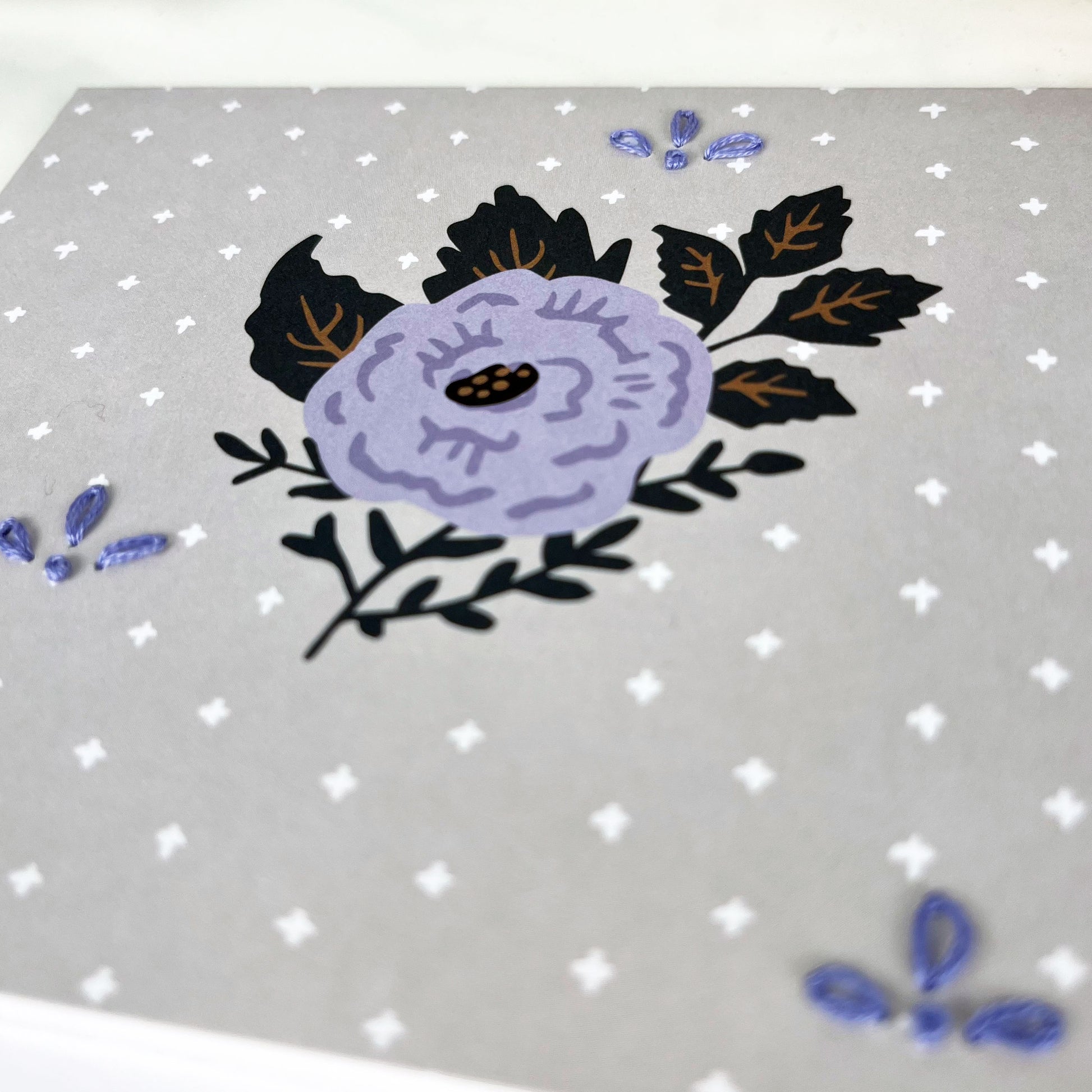 gray and purple floral greeting card embellished with small hand embroidered flowers on a white counter