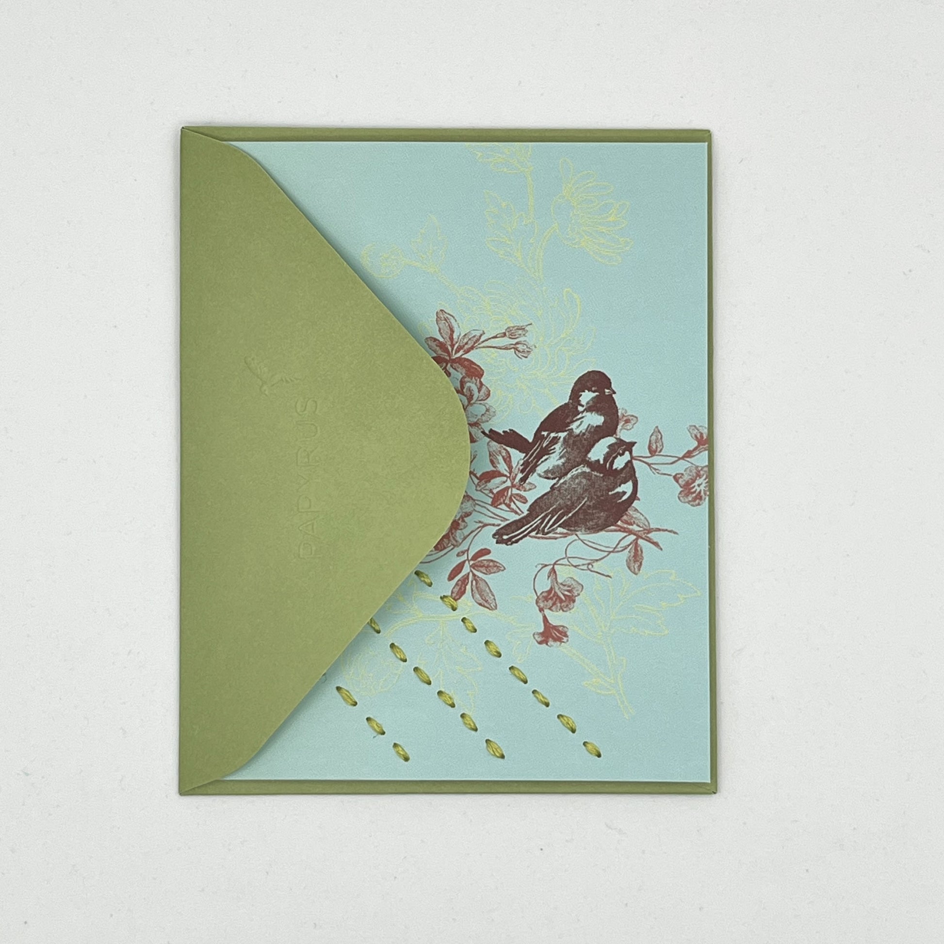 a blue greeting card laying flat, tucked in the flap of a green envelope with a sparrow and flowers printed on it in reds and chartreuse, hand stitched over with 3 rows of running stitches in chartreuse, diagonally over the bottom left corner