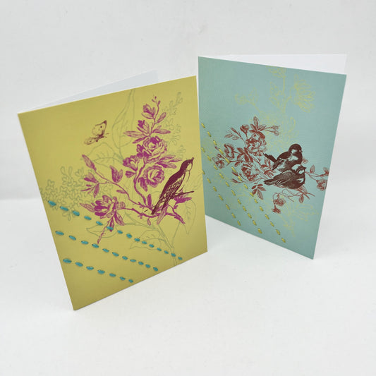 two greeting cards, one chartreuse and one robin's egg blue, standing upright, with a sparrow and flowers printed on it in brick red and fuchsia, hand stitched over with 3 rows of running stitches, diagonally over the bottom left corner