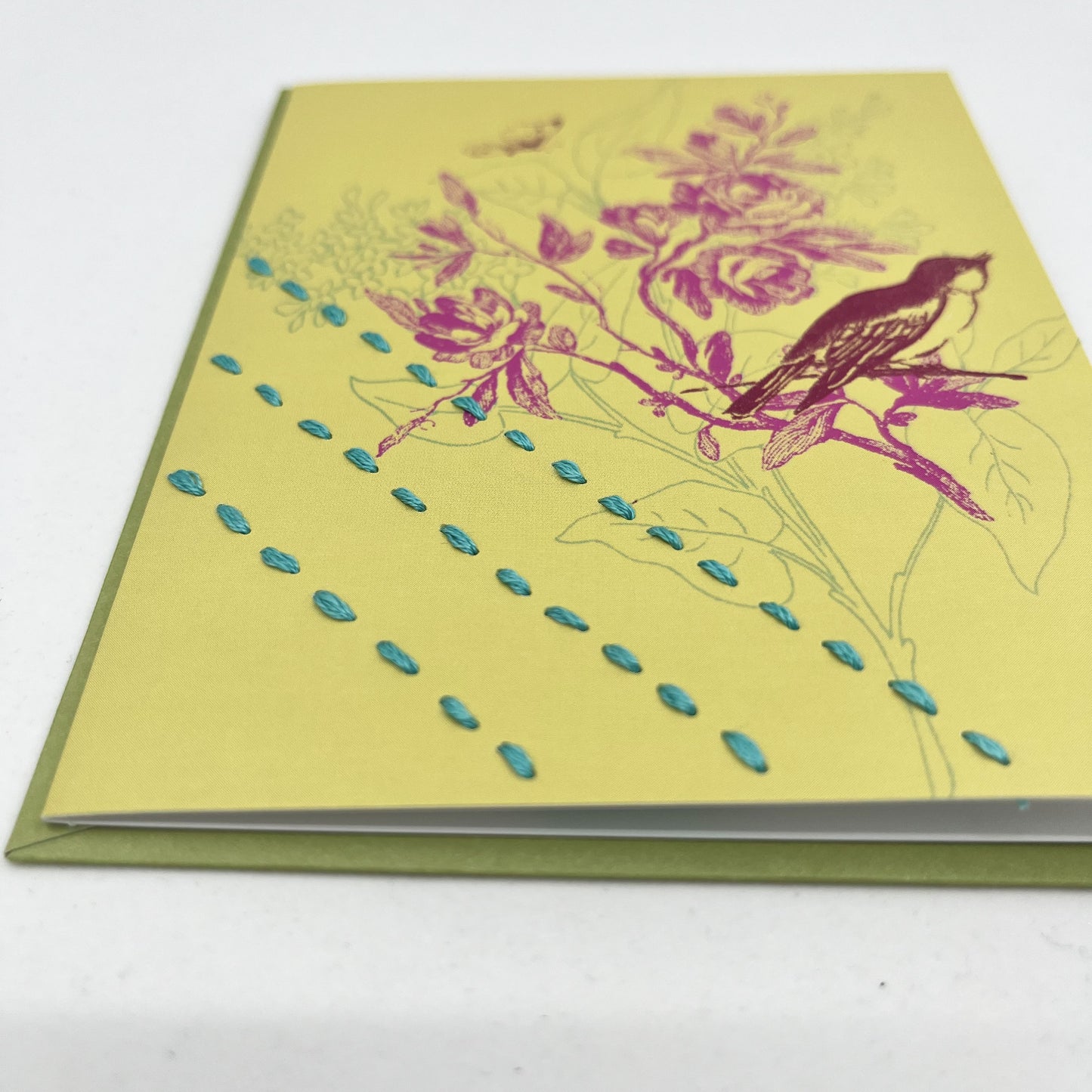close up angled view of a chartreuse greeting card with a sparrow and flowers printed on it in brick red and fuchsia, hand stitched over with 3 rows of running stitches in blue, diagonally over the bottom left corner