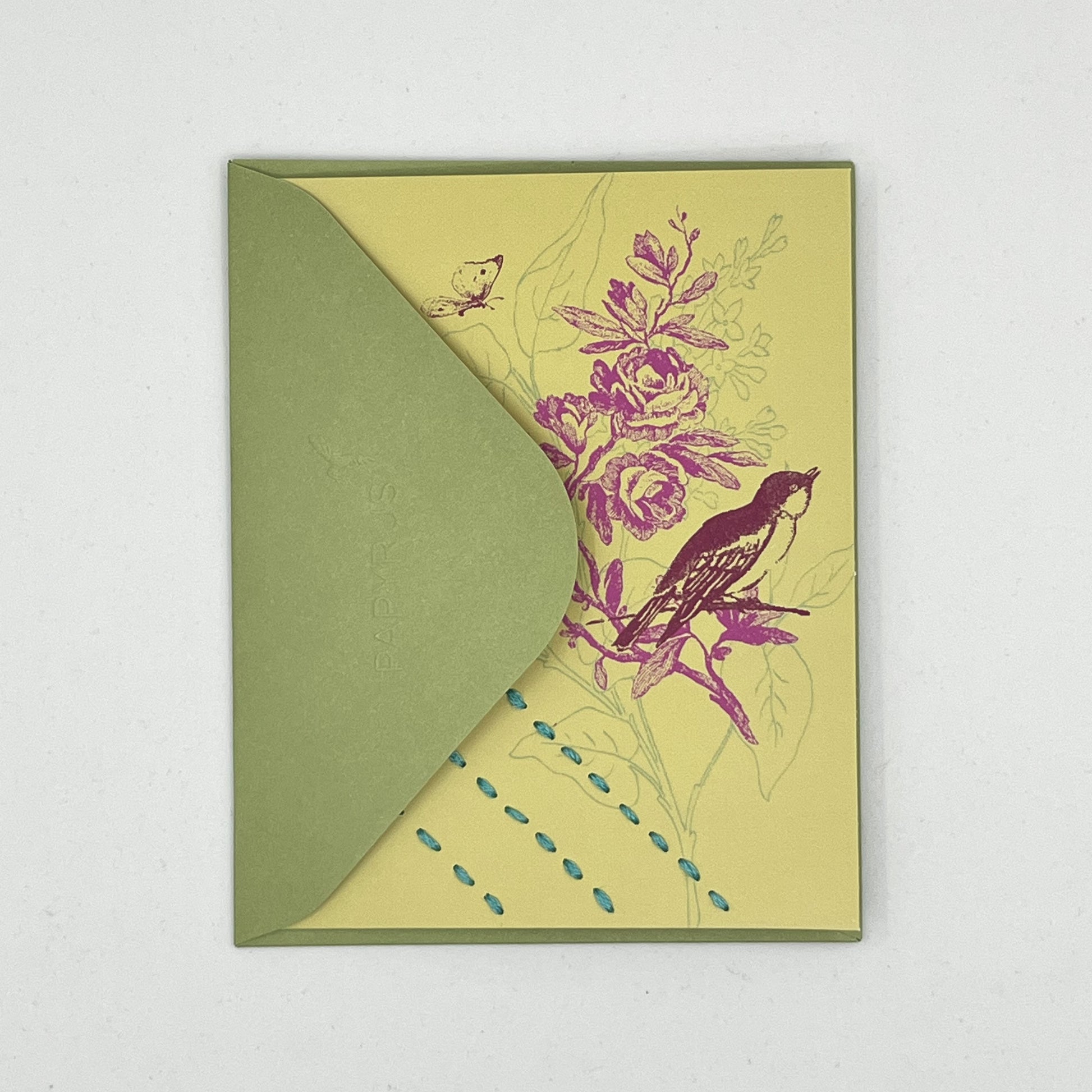 a chartreuse greeting card laying flat, tucked into the flap of a green envelope, with a sparrow and flowers printed on it in brick red and fuchsia, hand stitched over with 3 rows of running stitches in blue, diagonally over the bottom left corner