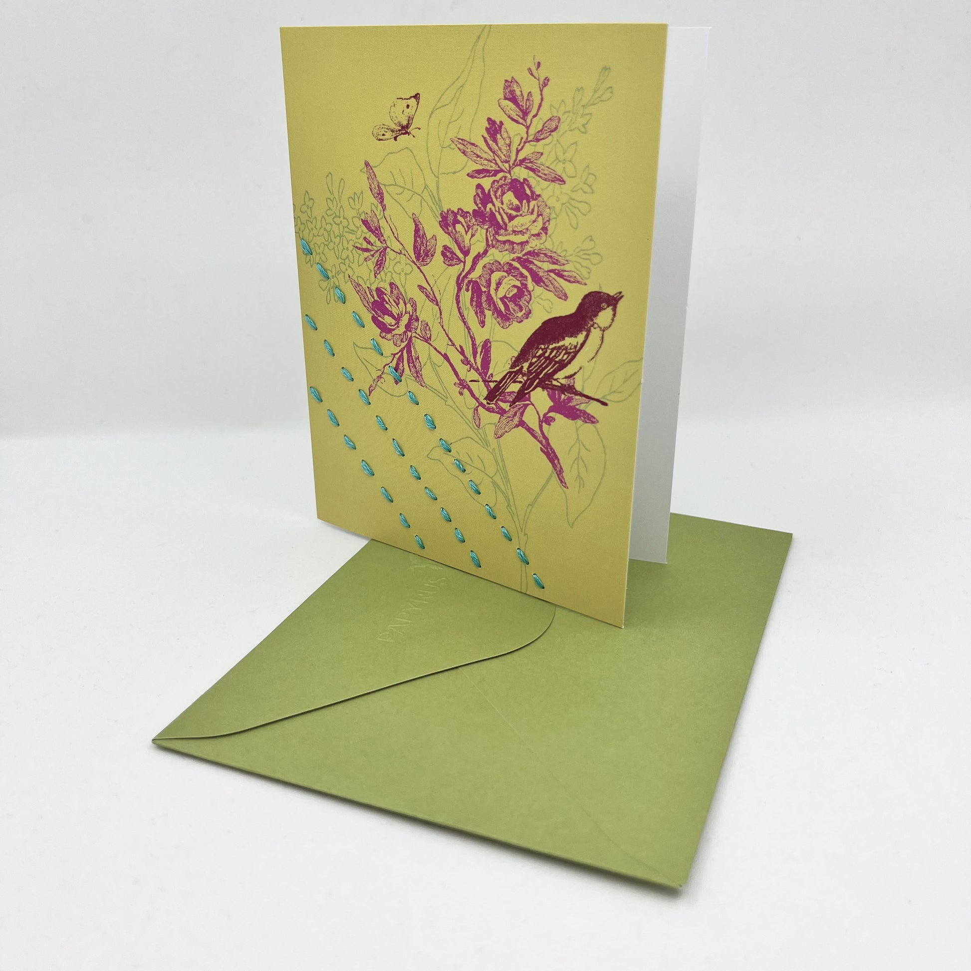 a chartreuse greeting card standing upright on a green envelope with a sparrow and flowers printed on it in brick red and fuchsia, hand stitched over with 3 rows of running stitches in blue, diagonally over the bottom left corner