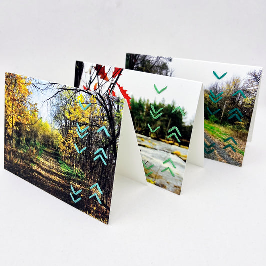 a group of three greeting cards standing up, depicting fall scenes of woods, trails, a river, hand stitched over with rows of unevenly spaced chevrons down the right side in aqua, seafoam green and teal