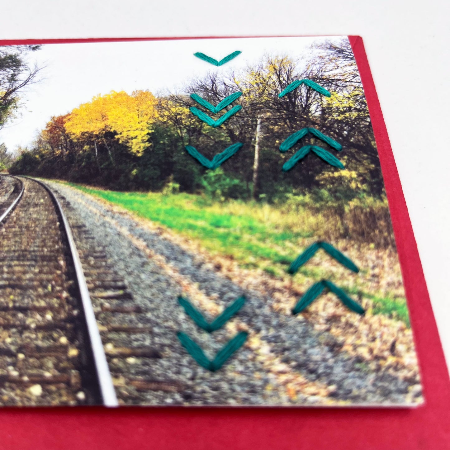 Greeting card flat on an envelope. Photo of a path through the woods in fall. Rows of aqua chevron stitches along right side card