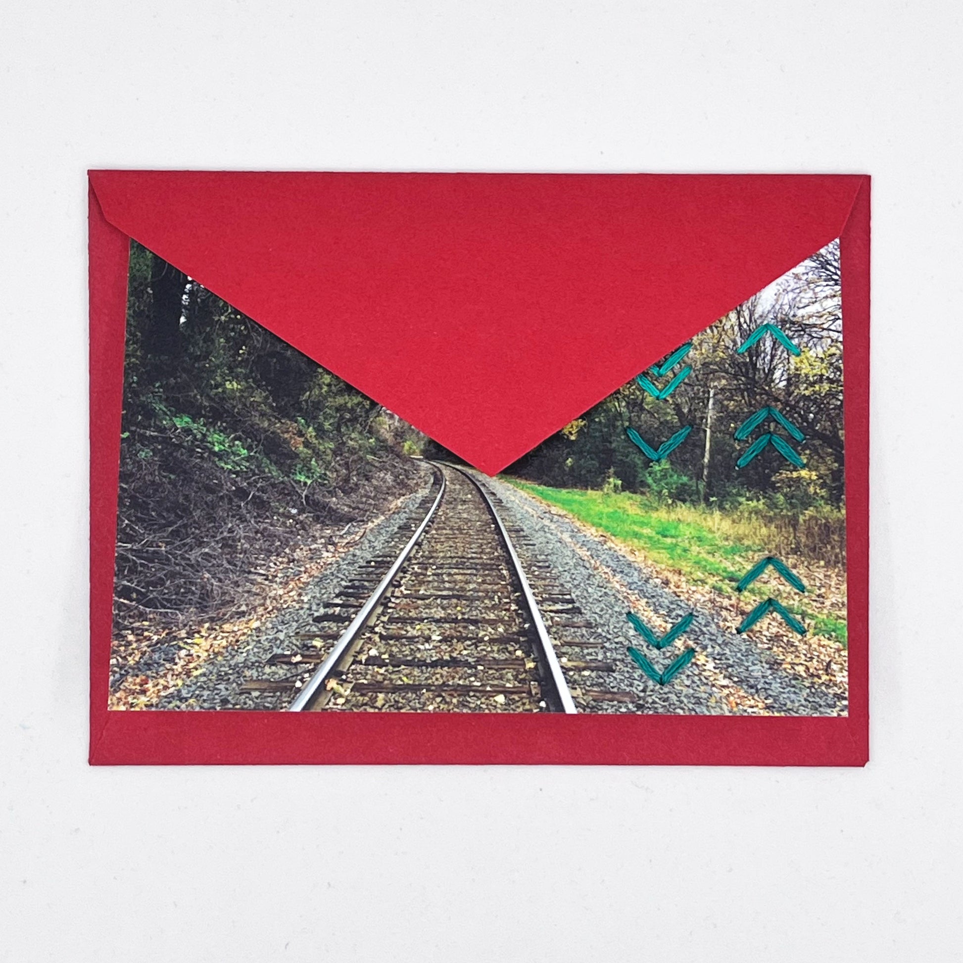 Greeting card flat on a red envelope, tucked under the flap. Photo of train tracks in fall. Rows of teal chevron stitches along right side card
