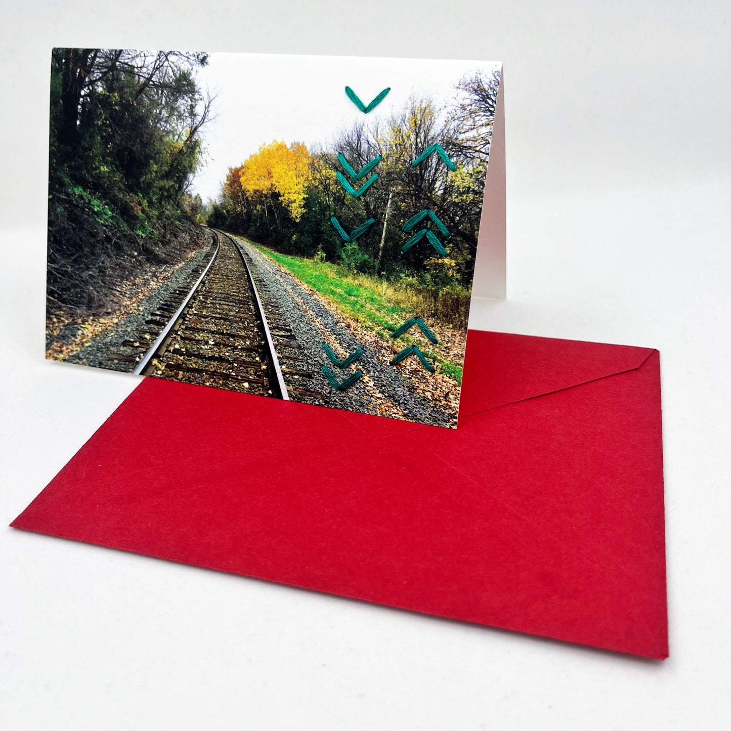 Greeting card standing upright on an envelope. Photo of train tracks in fall. Rows of teal chevron stitches along right side card