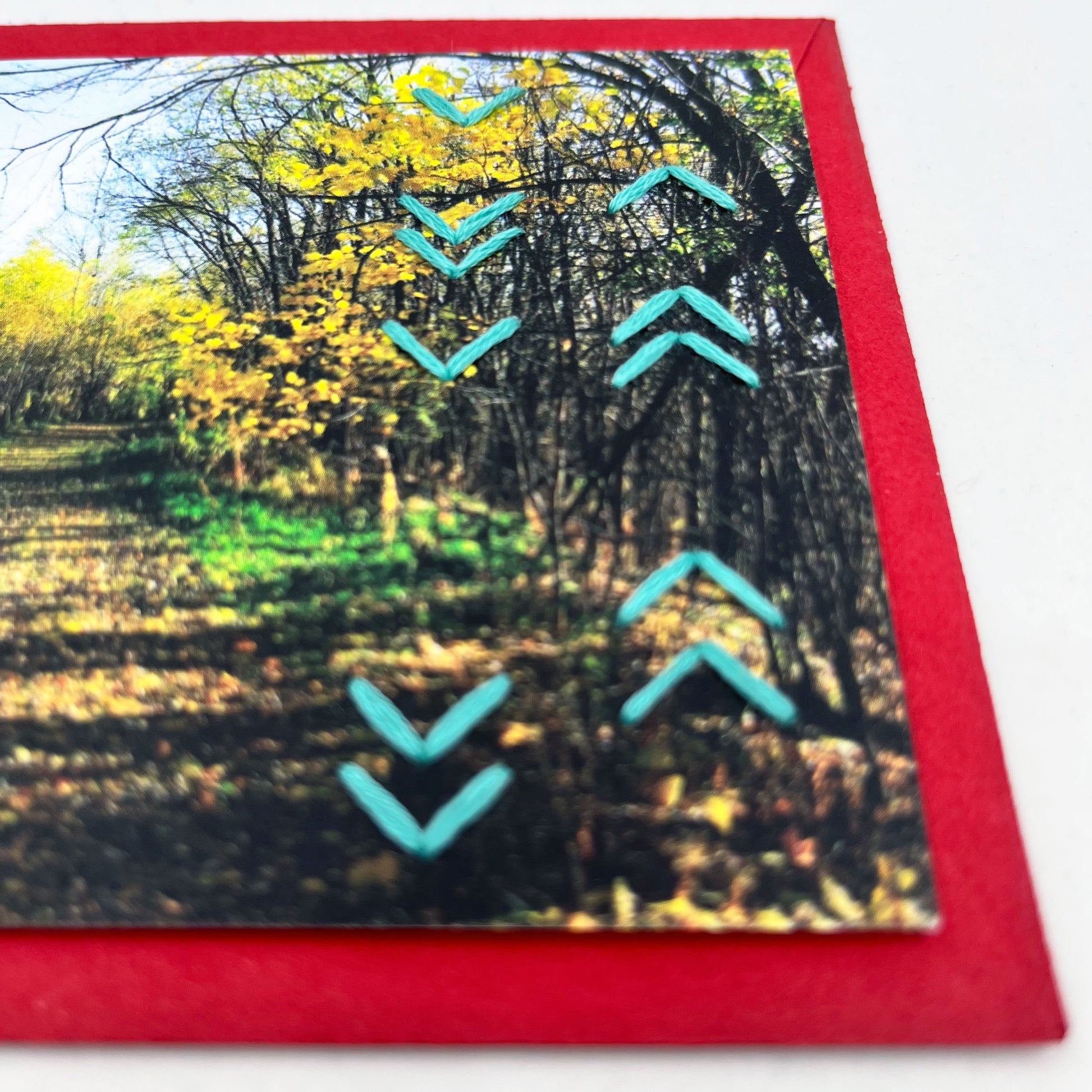 Greeting card flat on an envelope. Photo of a path through the woods in fall. Rows of aqua chevron stitches along right side card