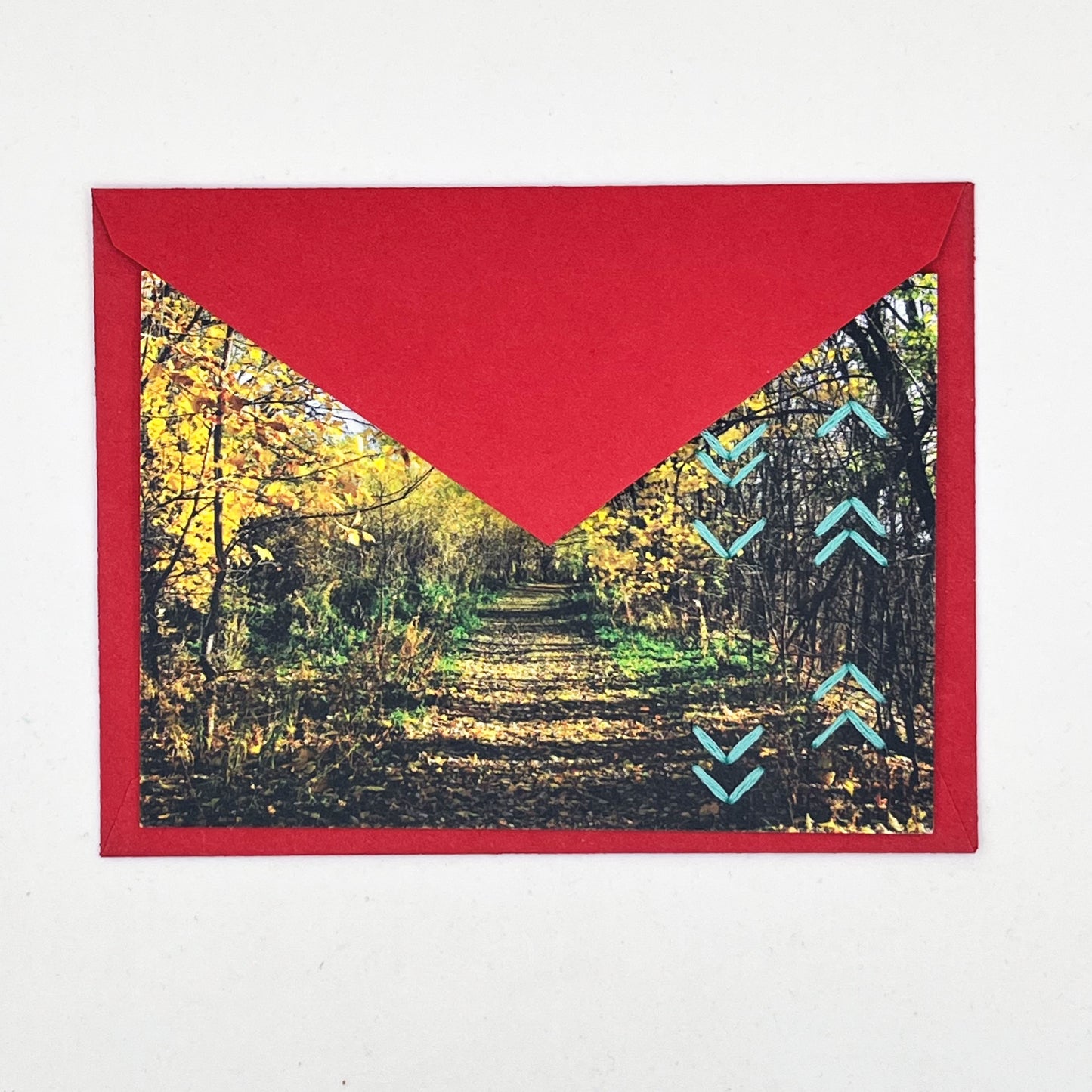 Greeting card flat on a red envelope, tucked under the flap. Photo of a path through the woods in fall. Rows of aqua chevron stitches along right side card