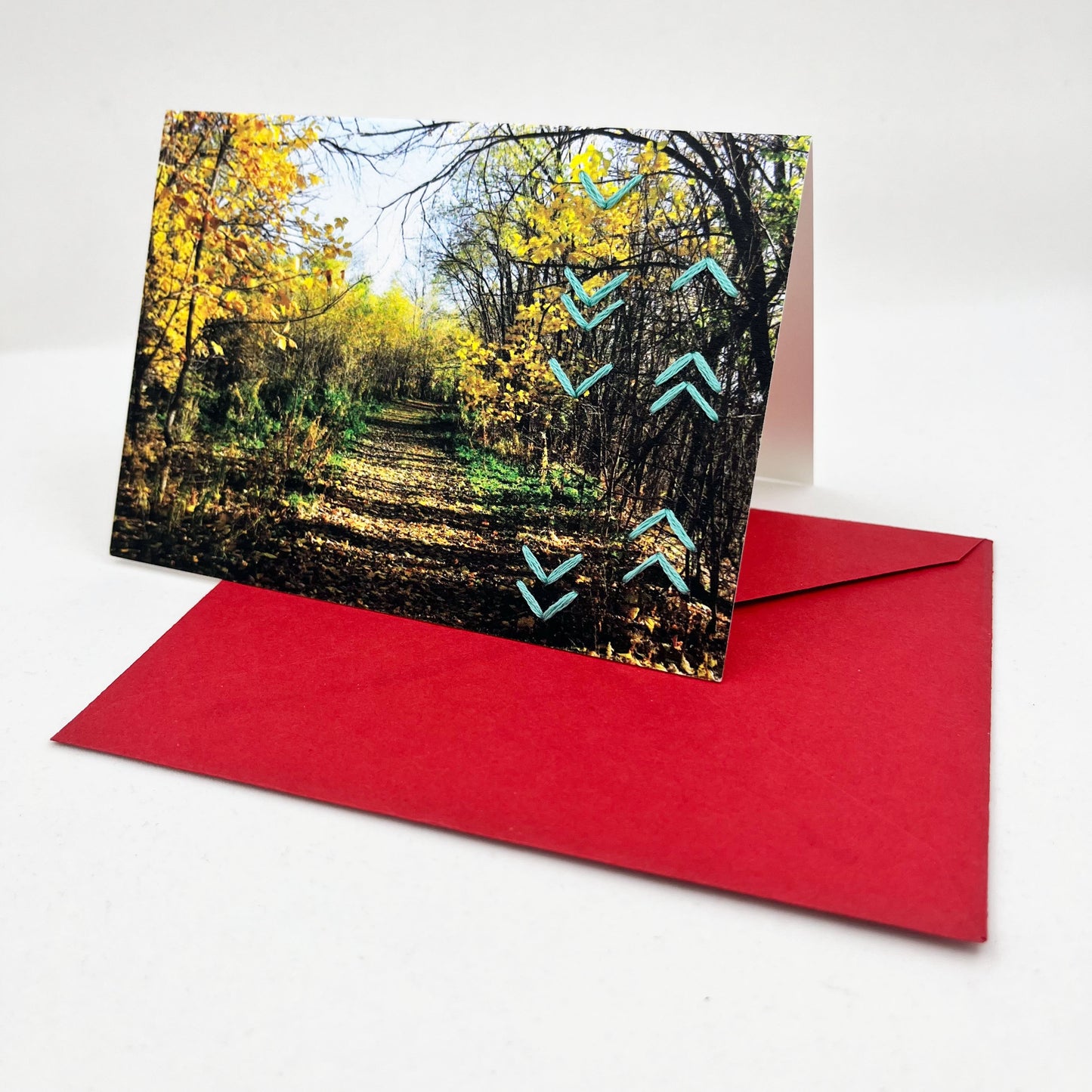 Greeting card standing upright on an envelope. Photo of a path through the woods in fall. Rows of aqua chevron stitches along right side card