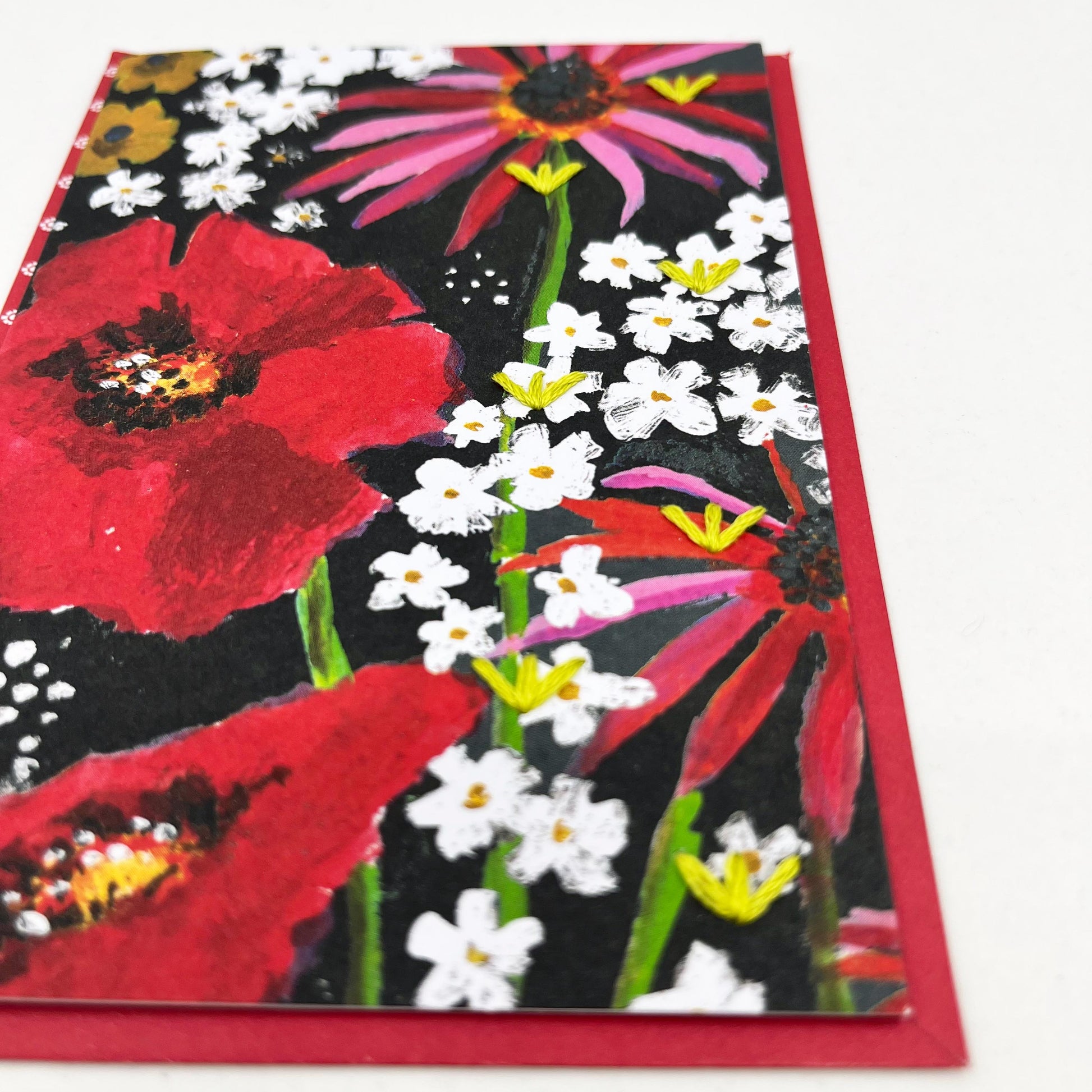 Greeting card standing flat on an envelope. artwork of painted flowers in red pink and white. Chartreuse stitches of sprout shapes in zig zag pattern on right side of card
