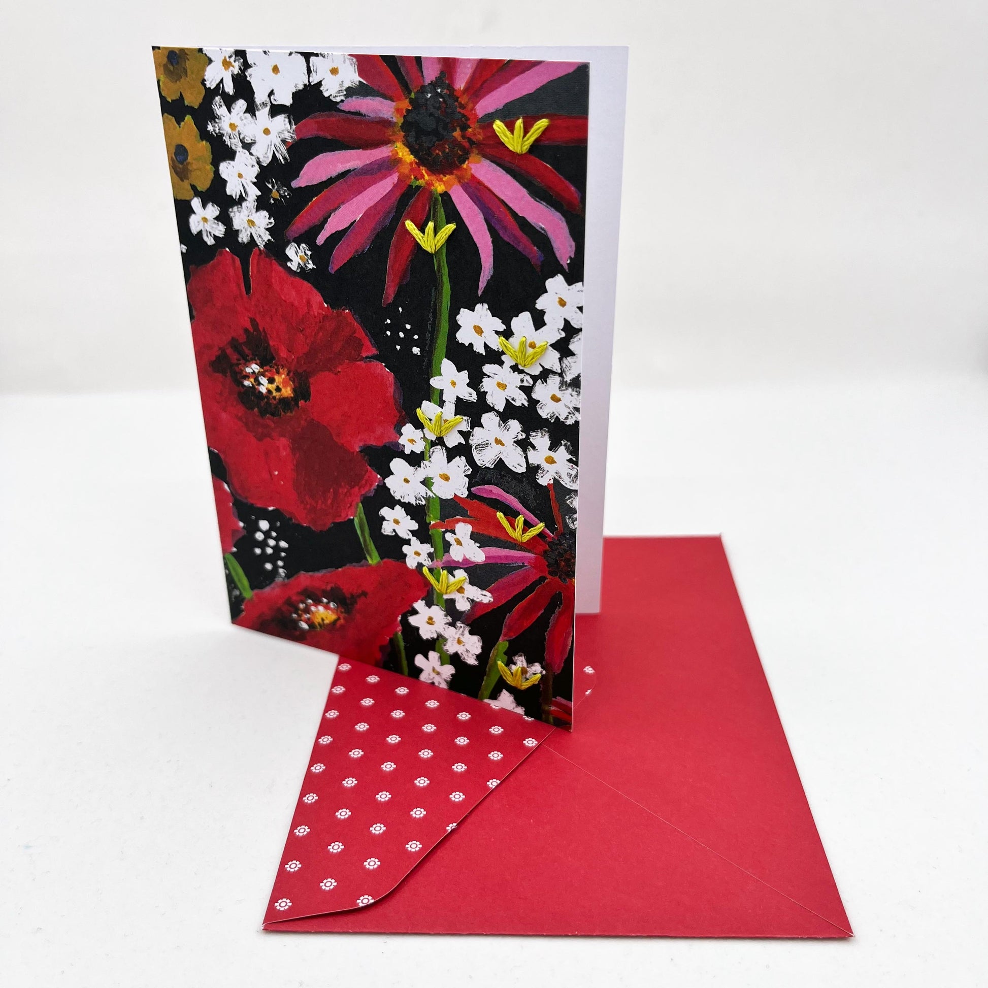 Greeting card standing upright on an envelope. artwork of painted flowers in red pink and white. Chartreuse stitches of sprout shapes in zig zag pattern on right side of card