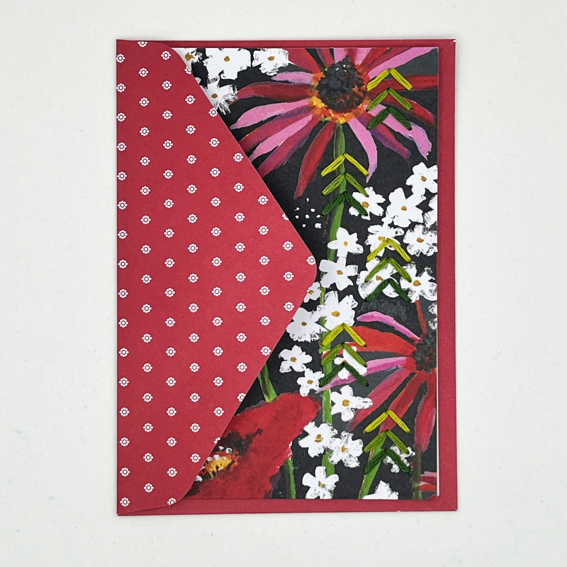Greeting card tucked into flap of an envelope. artwork of painted flowers in red pink and white. Green stitches of groups of 3 chevrons to create abstract pine tree shape