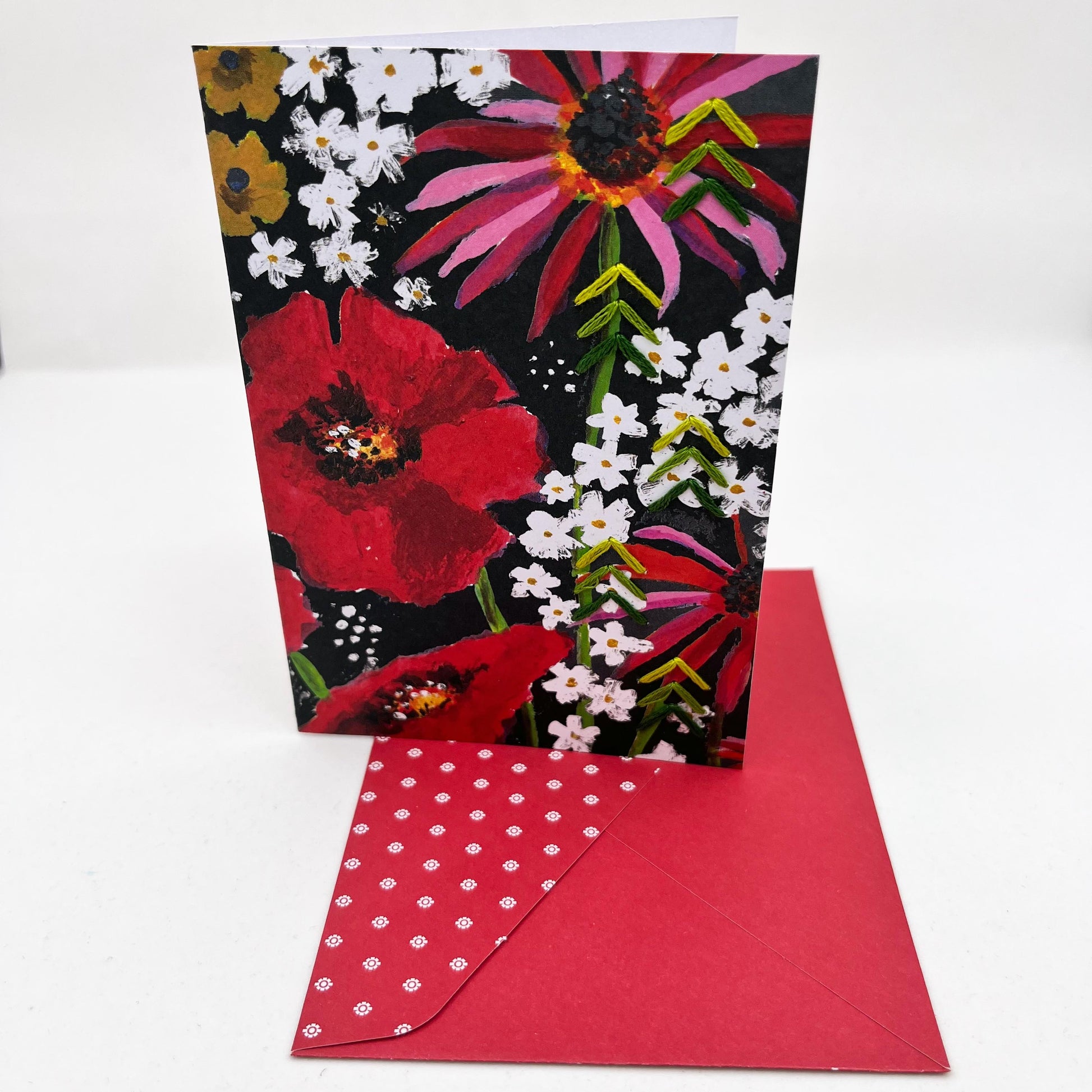 Greeting card standing upright on an envelope. artwork of painted flowers in red pink and white. Green stitches of groups of 3 chevrons to create abstract pine tree shape