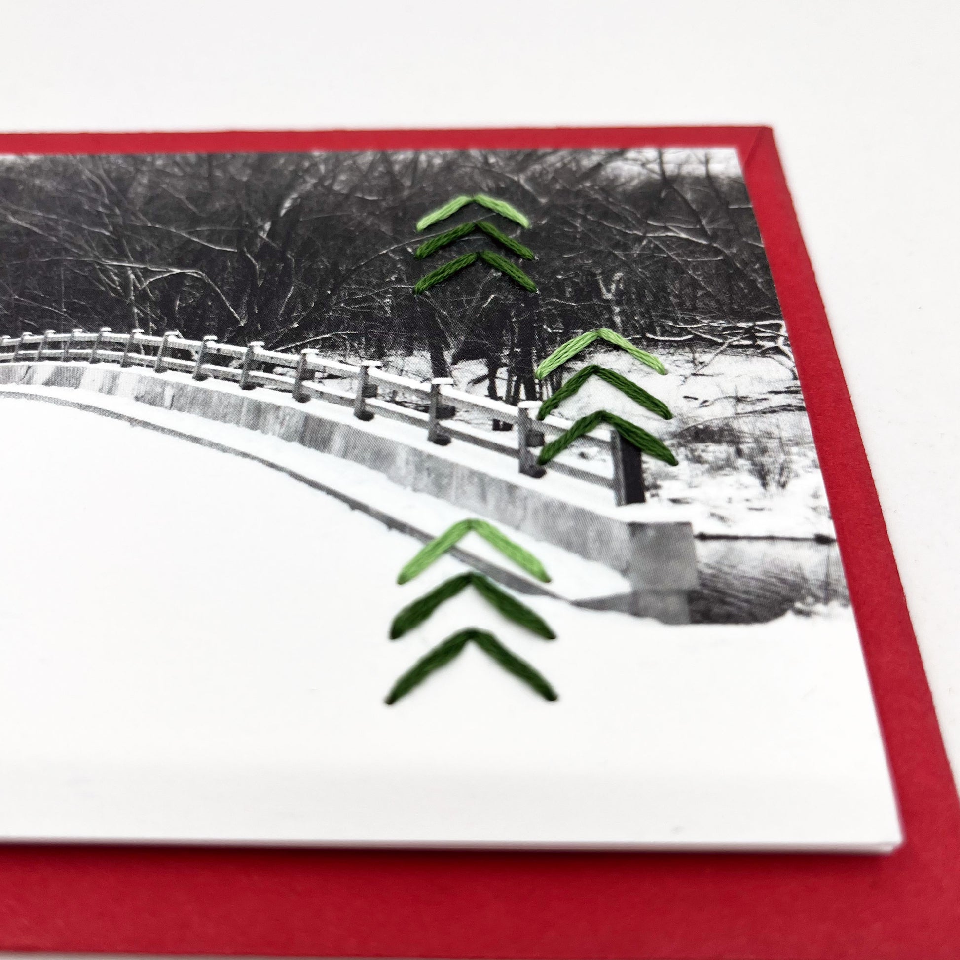 Greeting card flat on an envelope. Photo of snow covered bridge. Green stitches of groups of 3 chevrons to create abstract pine tree shape