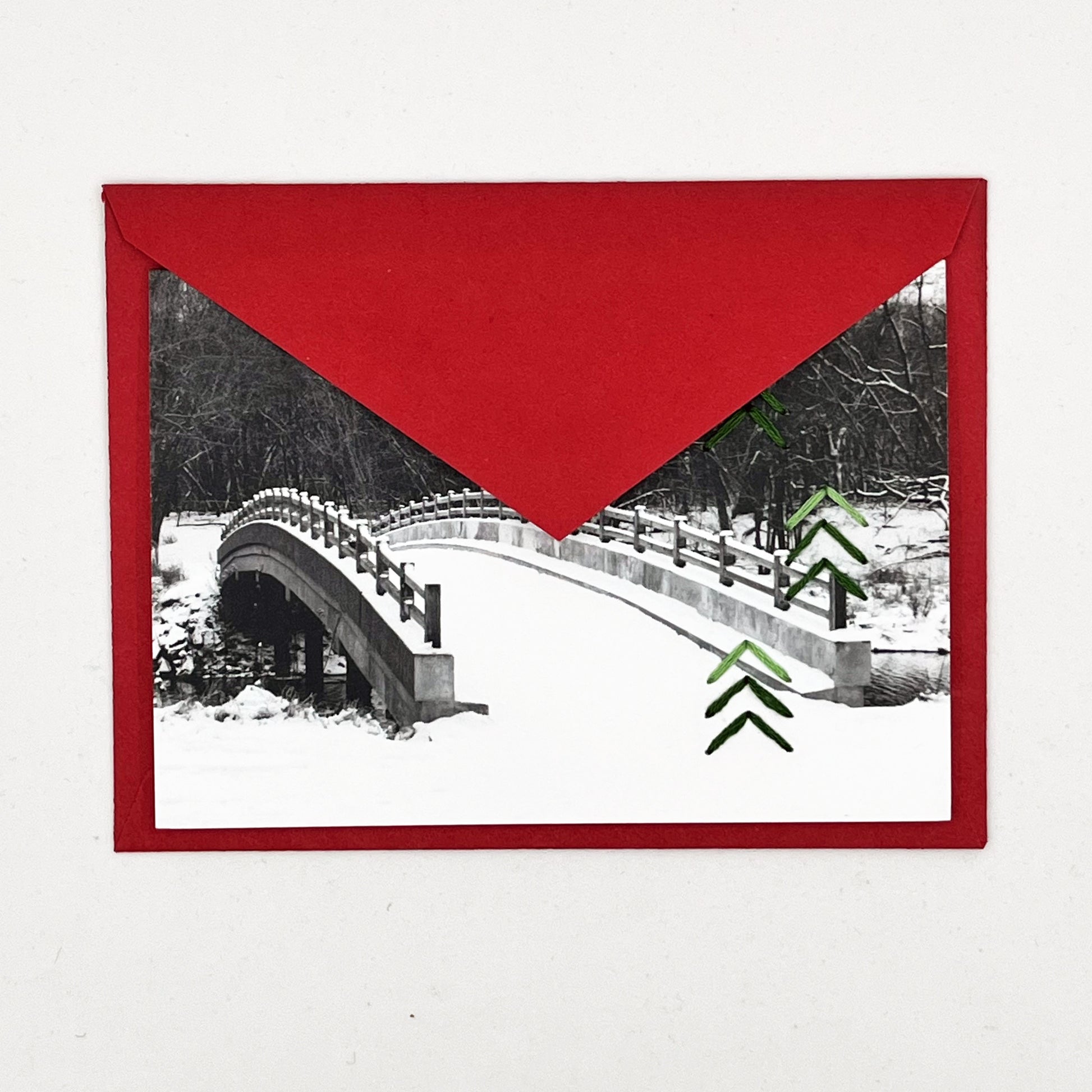 Greeting card flat on a red envelope, tucked under the flap. Photo of snow covered bridge. Green stitches of groups of 3 chevrons to create abstract pine tree shape