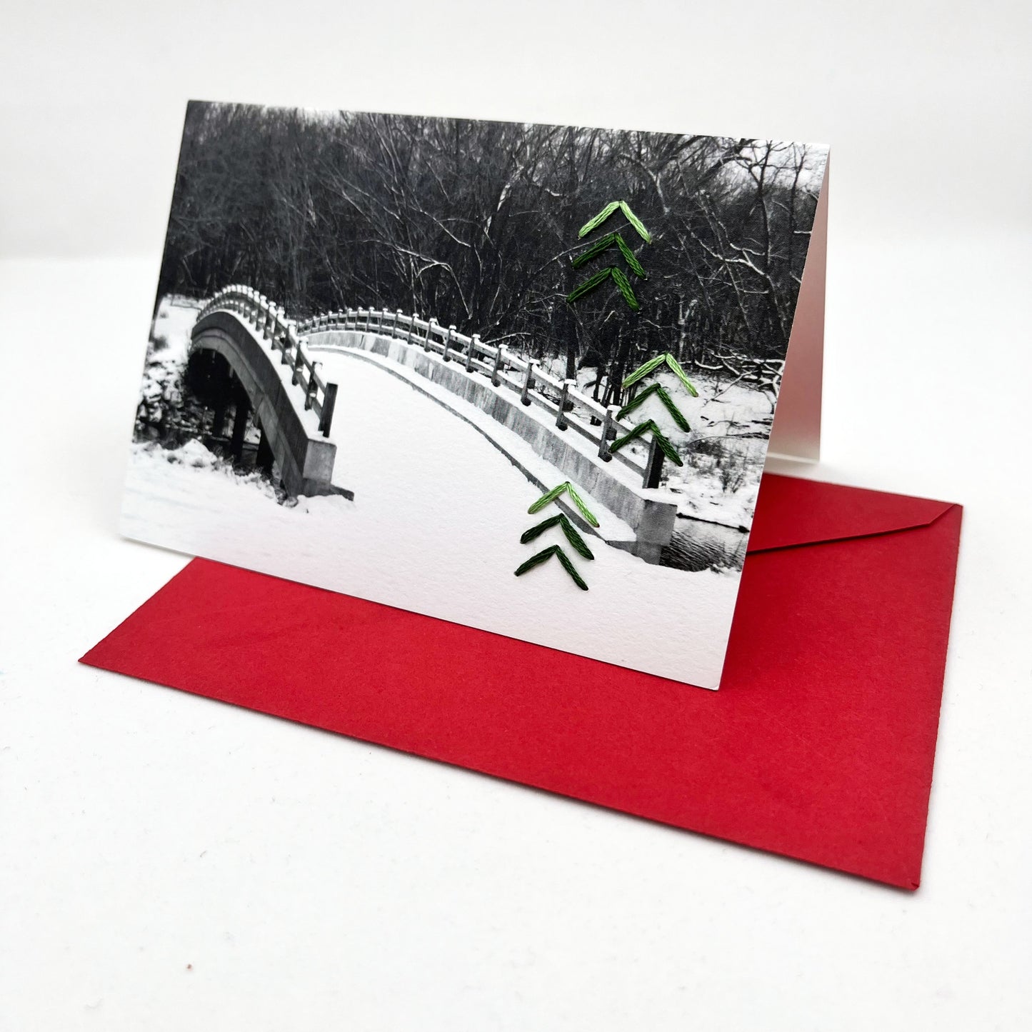 Greeting card standing upright on an envelope. Photo of snow covered bridge. Green stitches of groups of 3 chevrons to create abstract pine tree shape