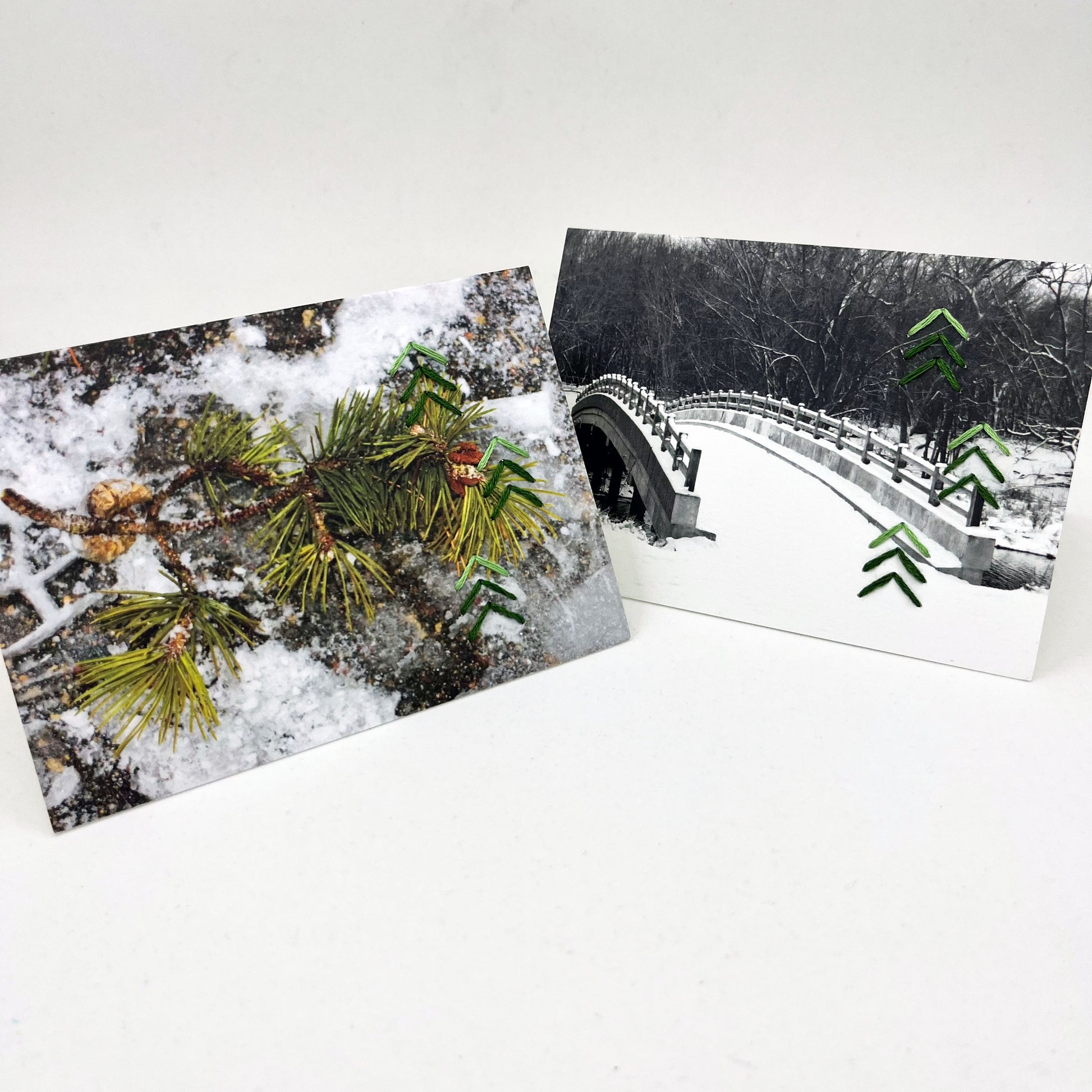 a group of two greeting cards standing up, depicting winter scenes of a snow covered bridge and a pine tree branch in snow, hand stitched over with groups of green chevrons resembling a pine tree down the right side