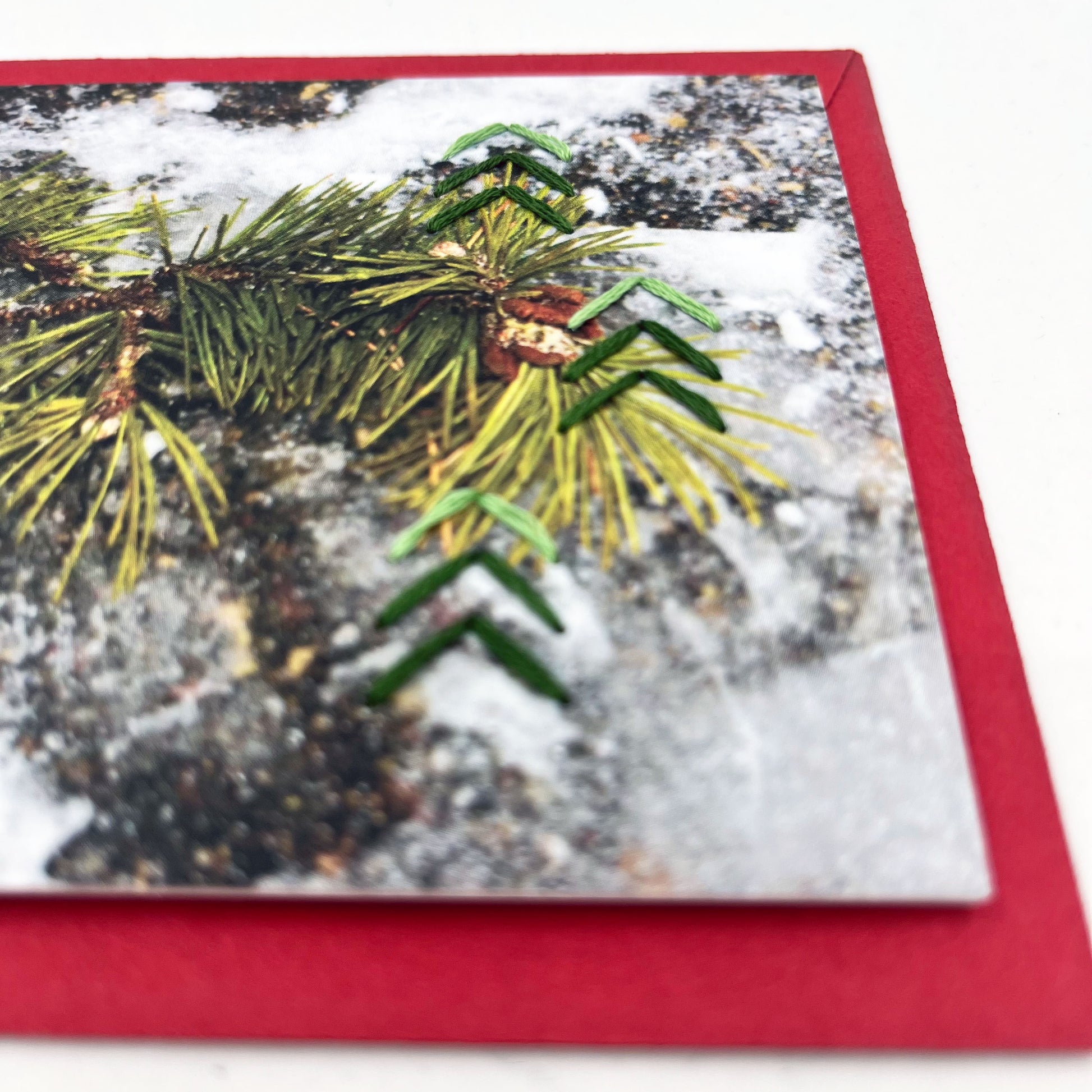 Greeting card flat on an envelope. Photo of pine tree branch in snow. Green stitches of groups of 3 chevrons to create abstract pine tree shape