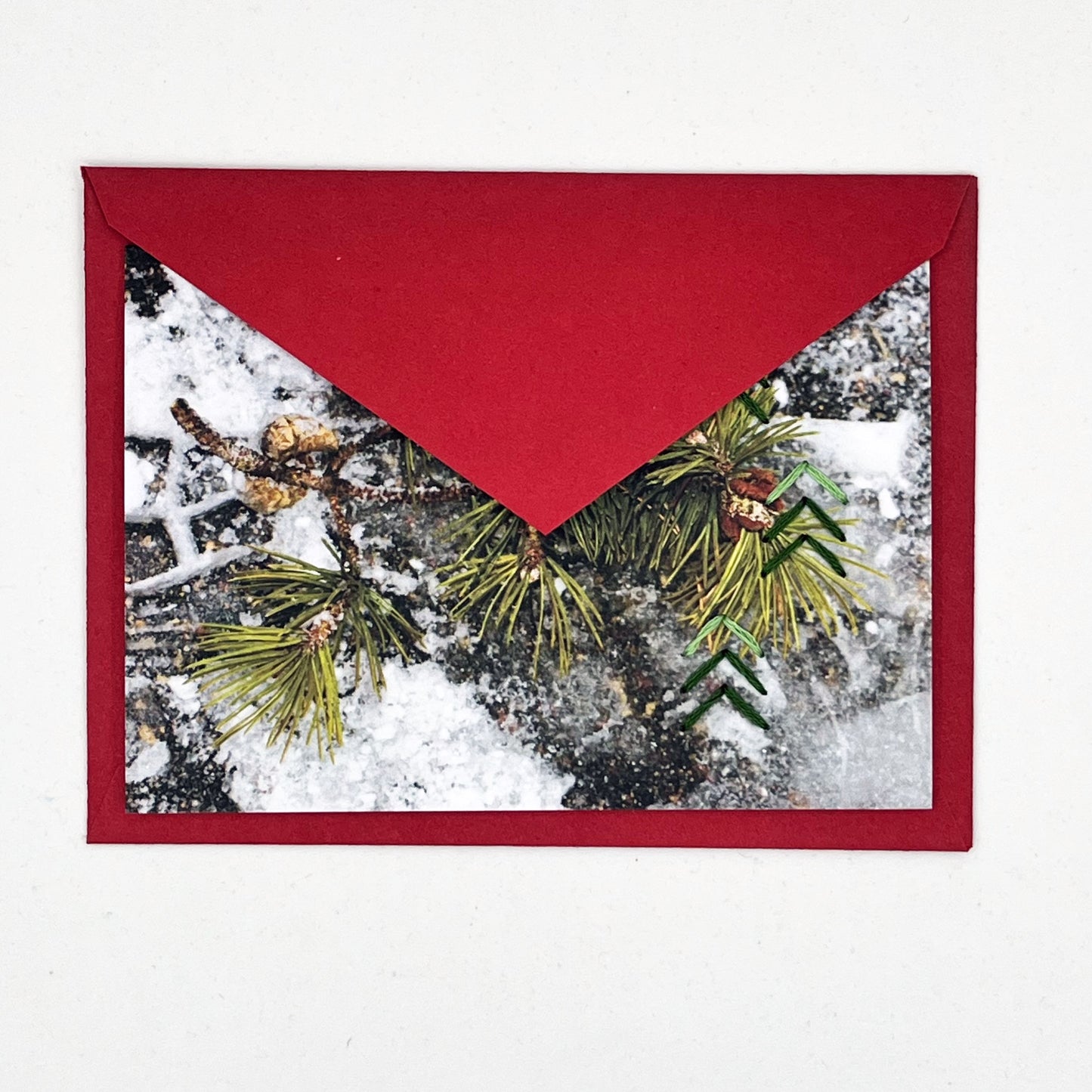 Greeting card flat on a red envelope, tucked under the flap. Photo of pine tree branch in snow. Green stitches of groups of 3 chevrons to create abstract pine tree shape