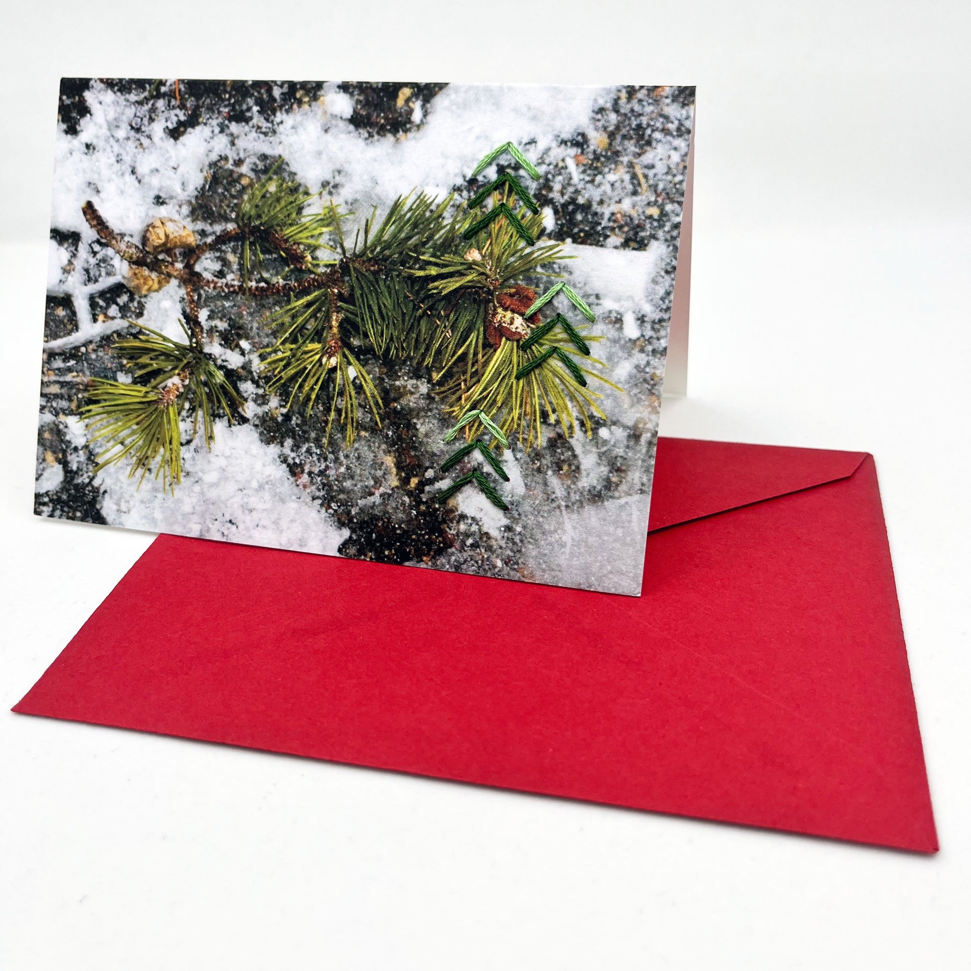Greeting card standing upright on an envelope. Photo of pine tree branch in snow. Green stitches of groups of 3 chevrons to create abstract pine tree shape