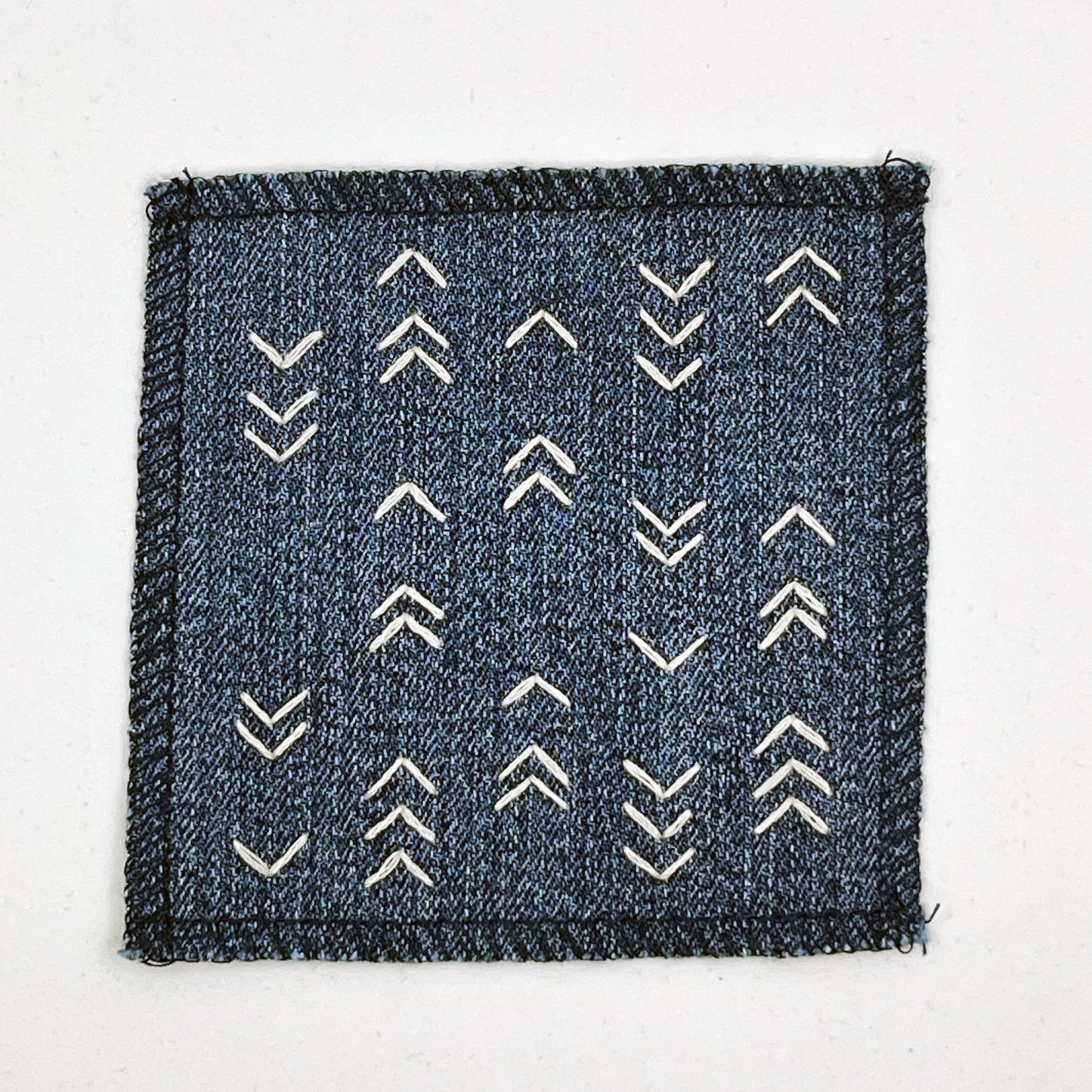  a square denim patch hand stitched in ivory with rows of unevenly spaced small chevrons facing different directions on a white background