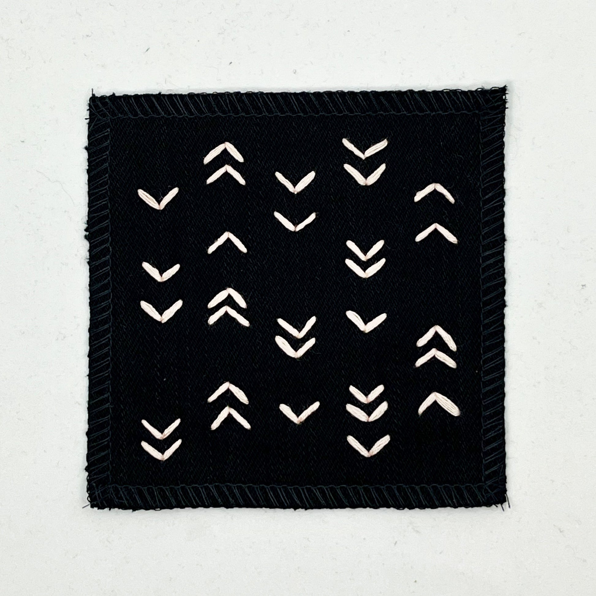 a square black denim patch hand stitched in peach with rows of unevenly spaced small chevrons facing different directions on a white background
