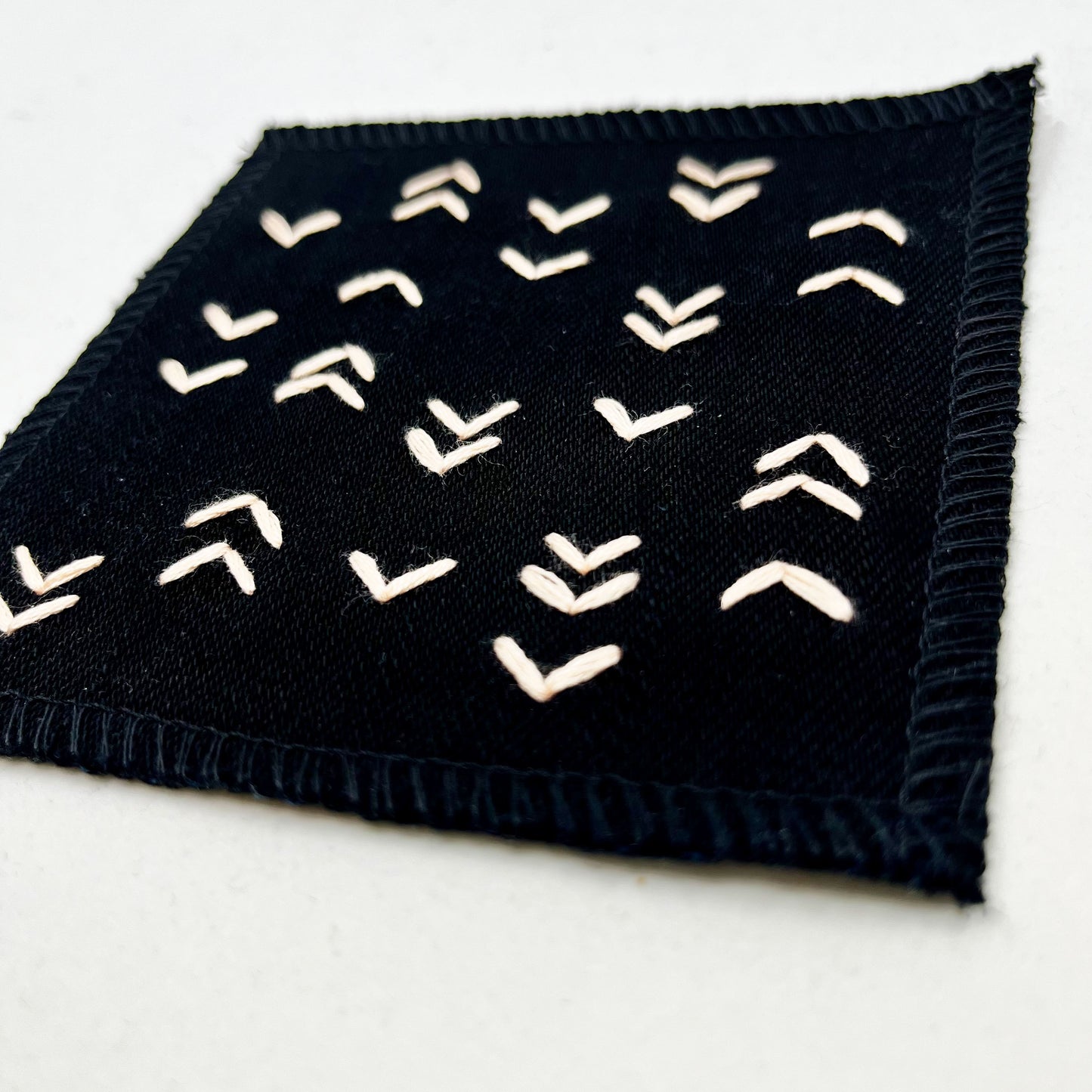 close up angled view of a square black denim patch hand stitched in peach with rows of unevenly spaced small chevrons facing different directions on a white background