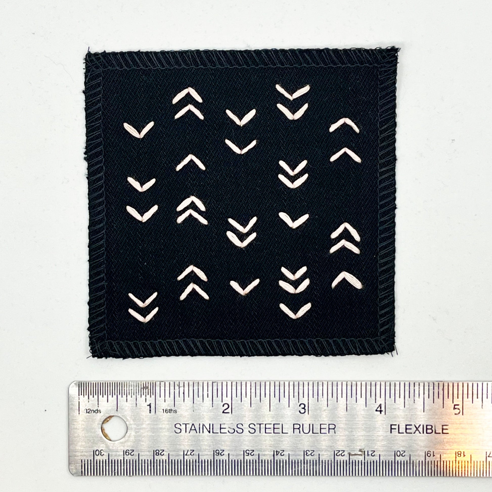 a square black denim patch hand stitched in peach with rows of unevenly spaced small chevrons facing different directions placed next to a metal ruler to show a width of four inches
