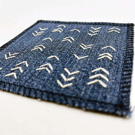 close up angled view of a square denim patch hand stitched in ivory with rows of unevenly spaced small chevrons facing different directions on a white background