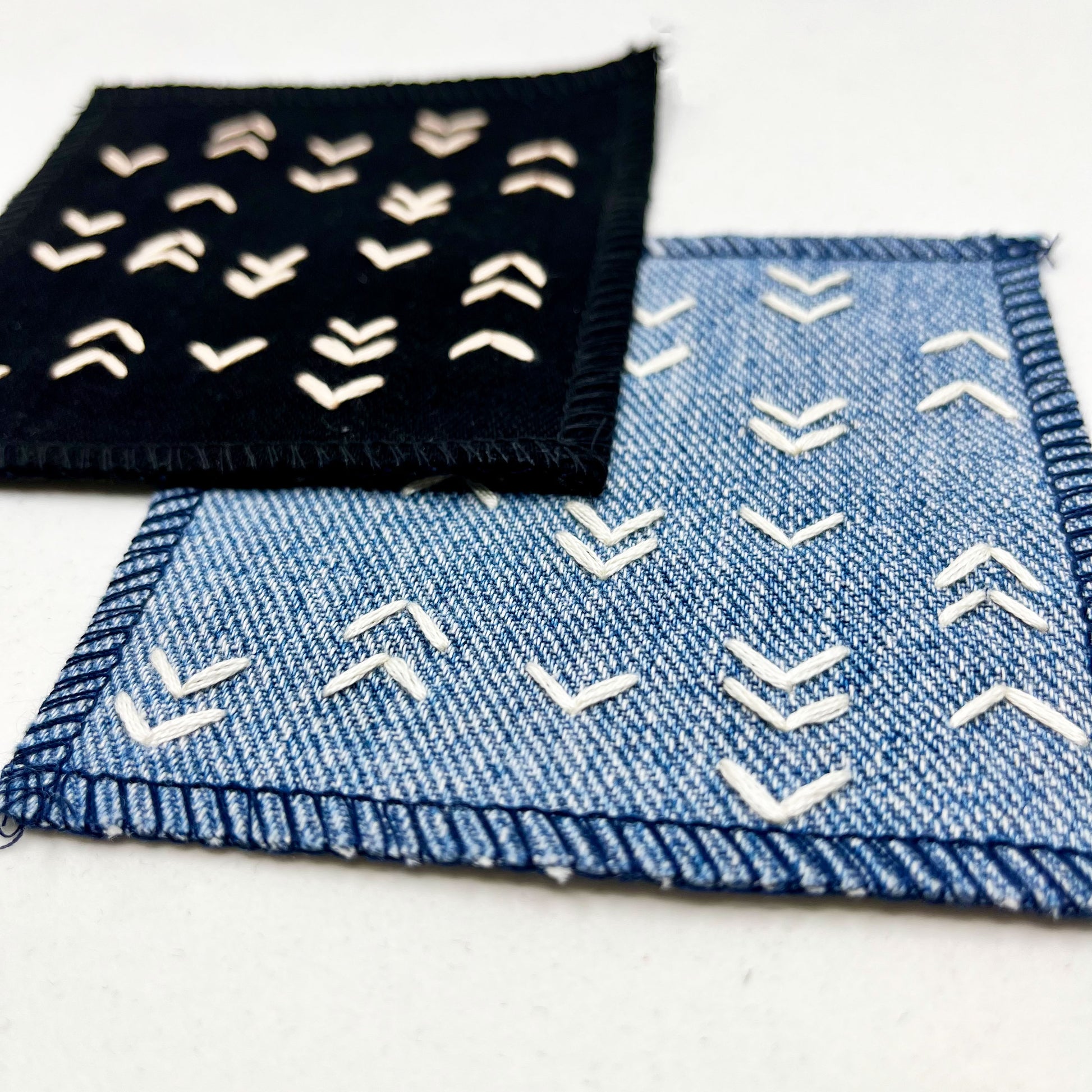 close up angled view of two square denim patches, one black one blue, hand stitched in peach and ivory respectively with rows of unevenly spaced small chevrons facing different directions on a white background