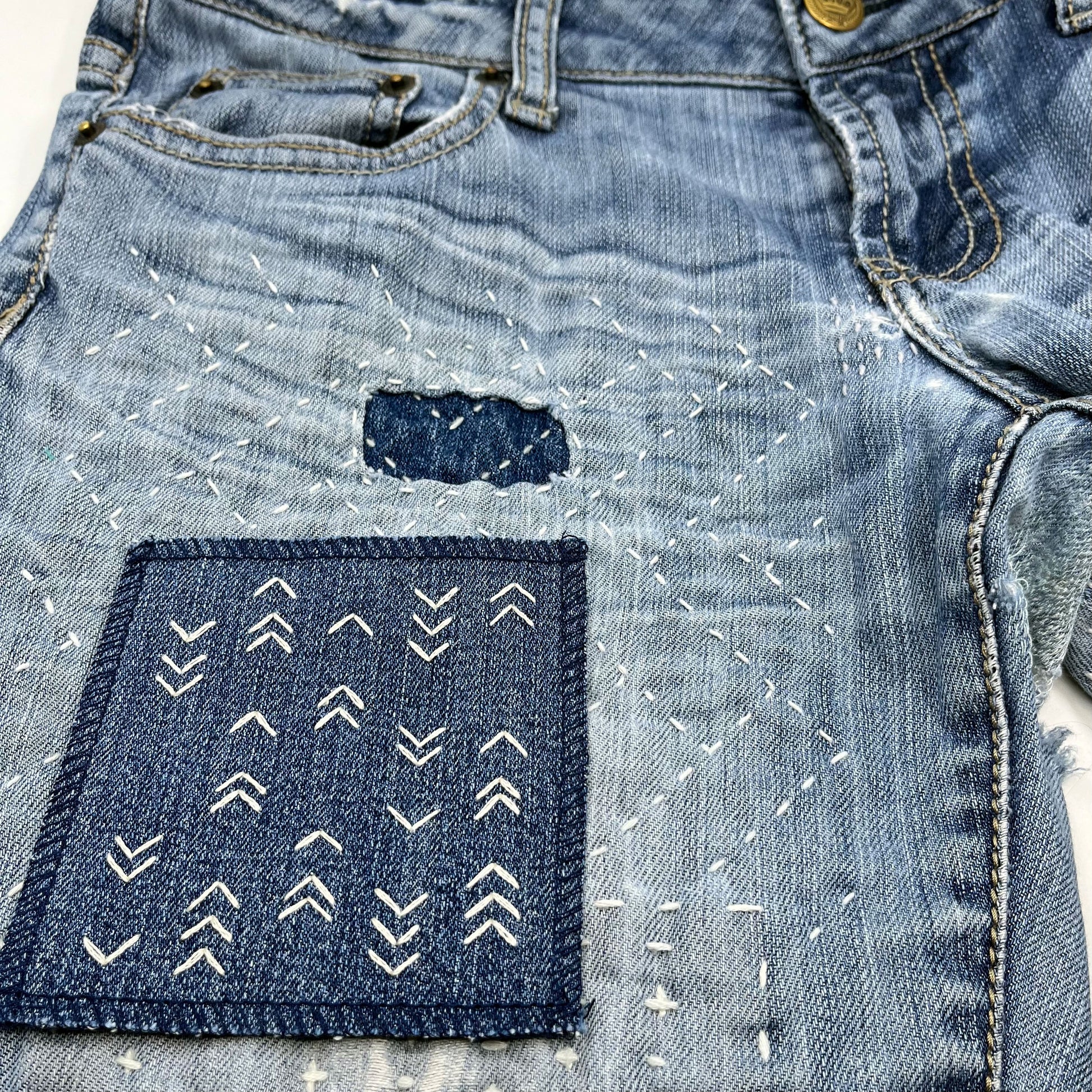 a square denim patch hand stitched in ivory with rows of unevenly spaced small chevrons facing different directions on pair of jeans heavily mended with sashiko stitching