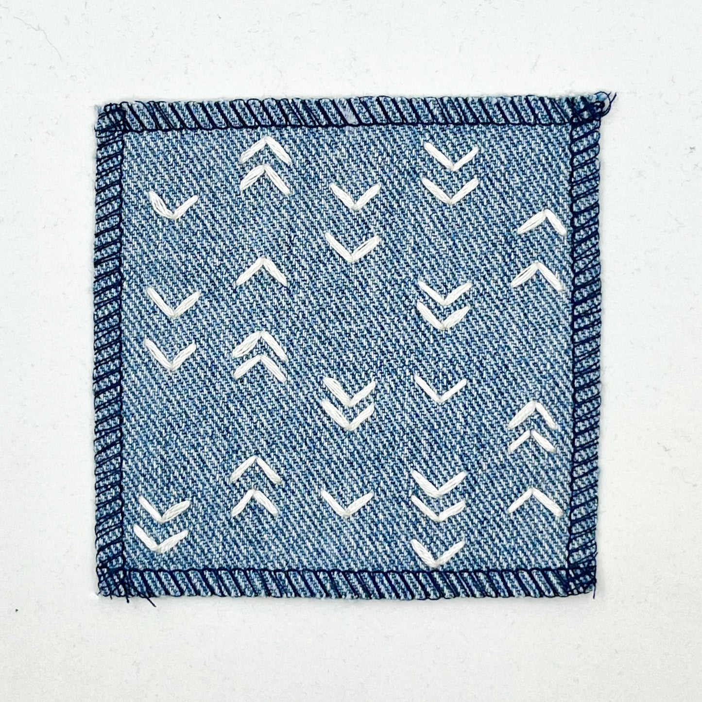a square denim patch hand stitched in ivory with rows of unevenly spaced small chevrons facing different directions on a white background
