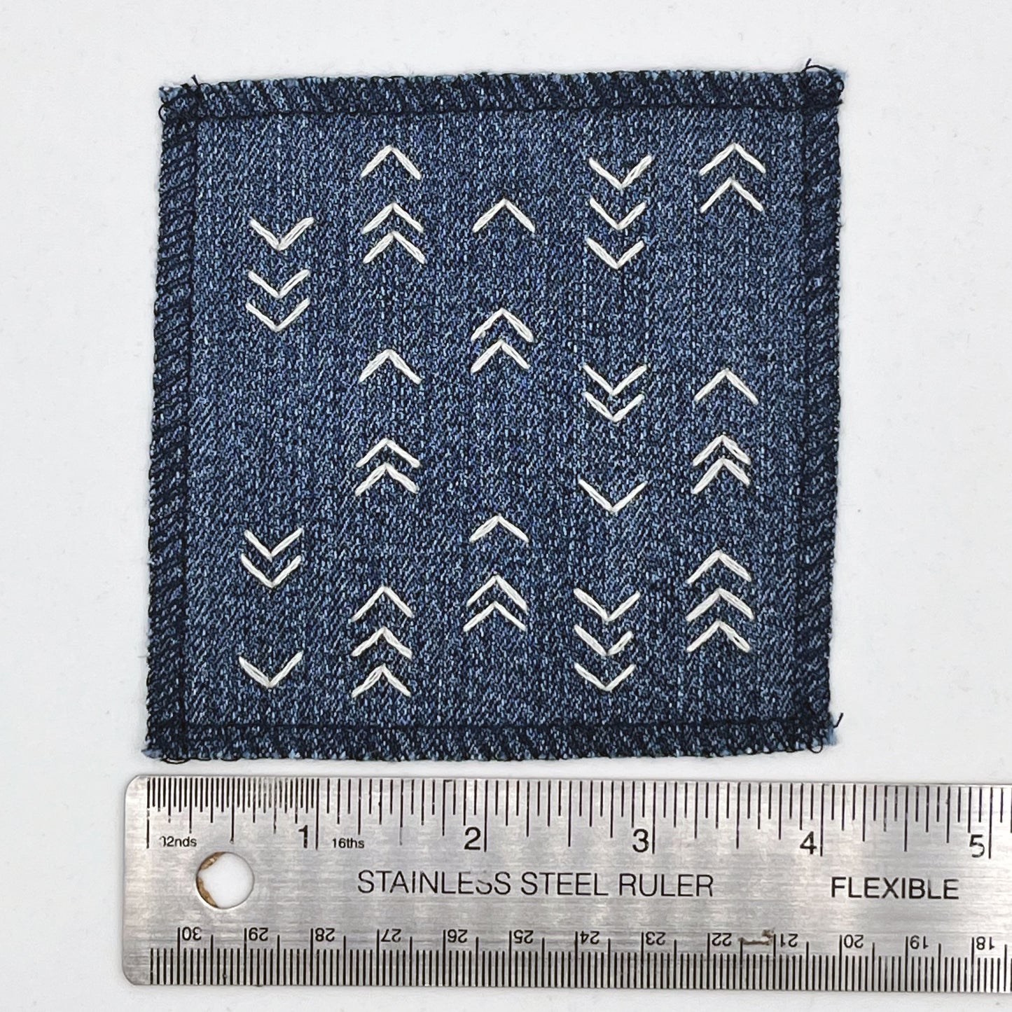 a square denim patch hand stitched in ivory with rows of unevenly spaced small chevrons facing different directions placed next to a metal ruler to show a width of four inches