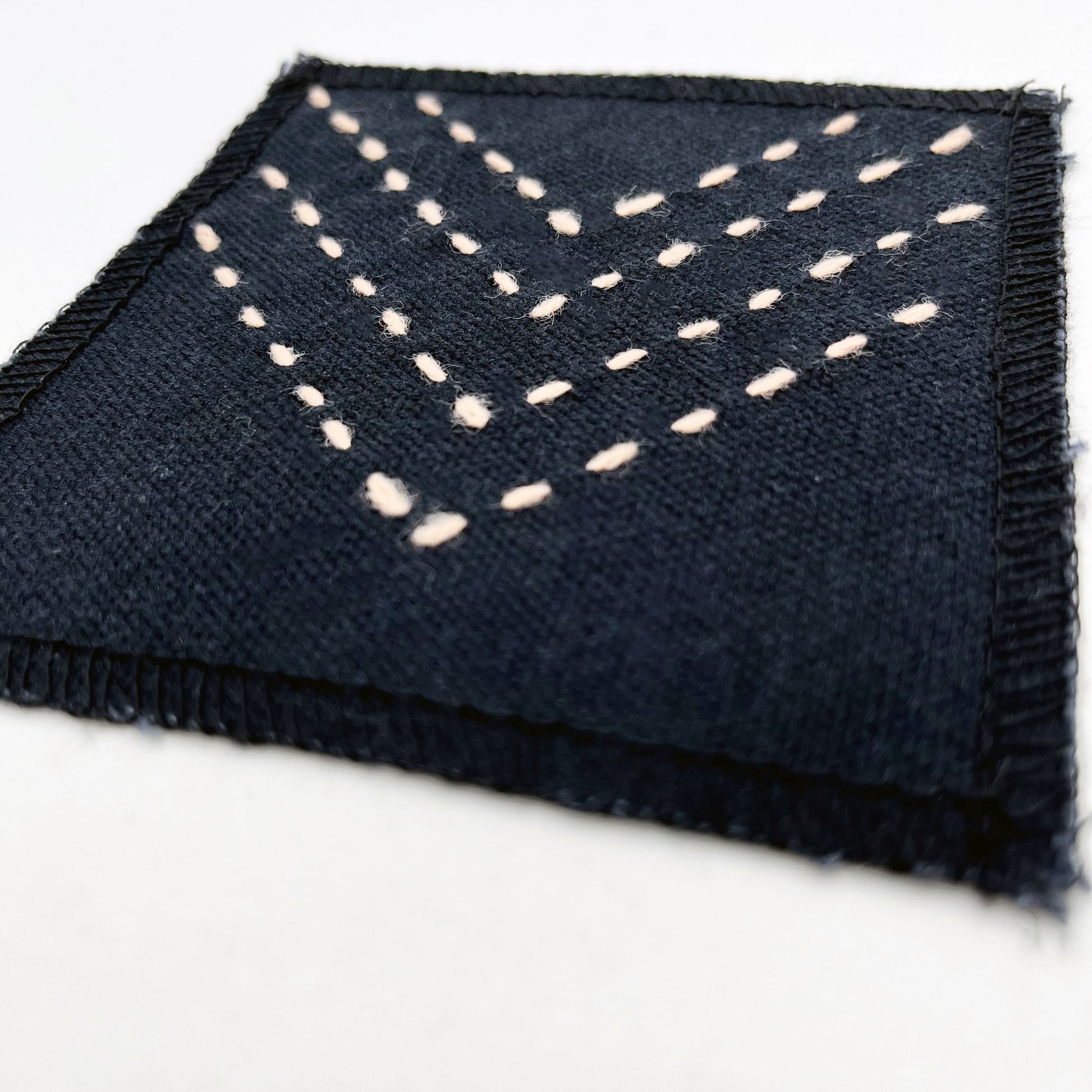 a close up angled view of a square patch made out of black canvas, handstitched with rows of peach running stitches in a chevron pattern, with overlocked edges, on a white background