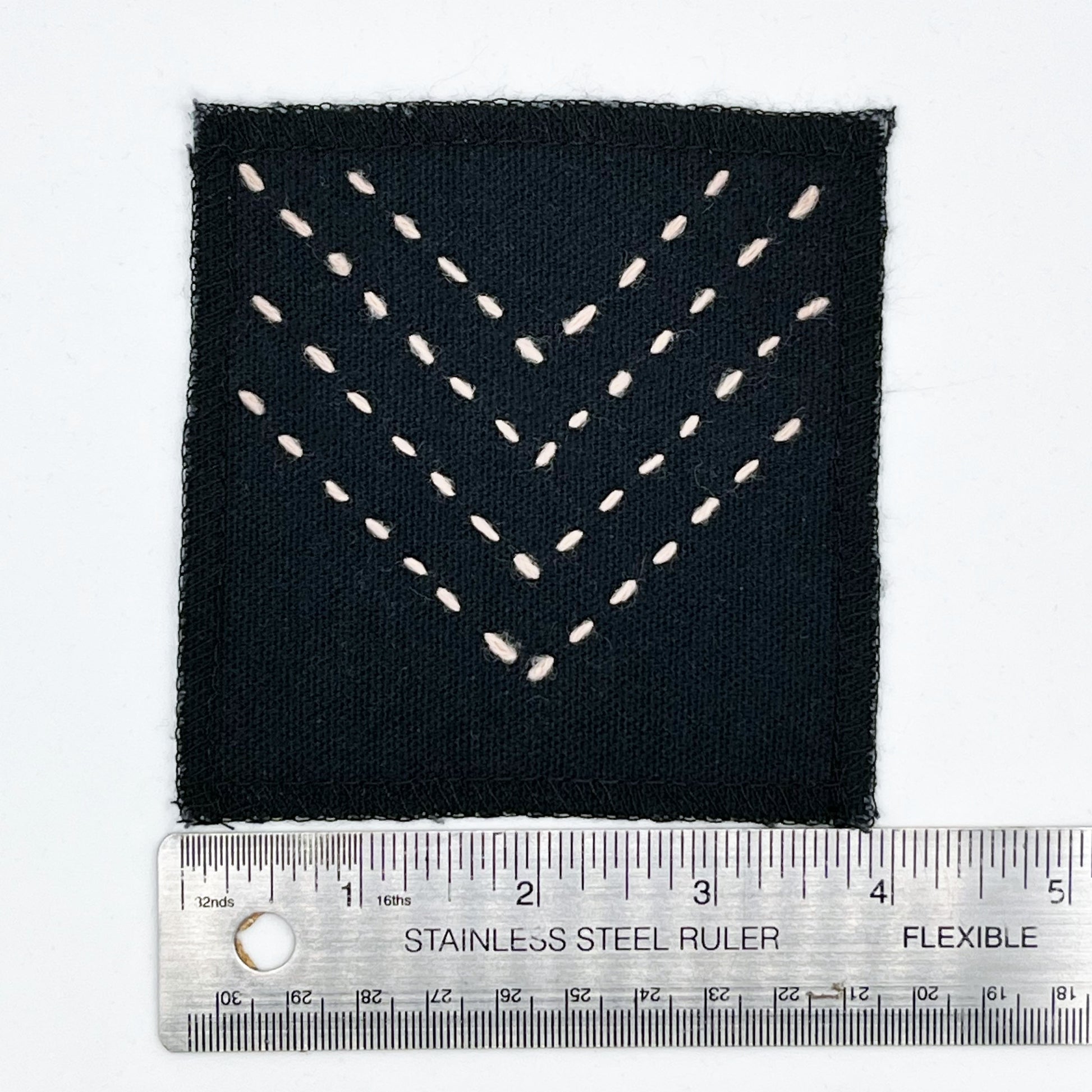 a square patch made out of black canvas, handstitched with rows of peach running stitches in a chevron pattern, with overlocked edges, next to a metal ruler to show a width of 4 inches, on a white background