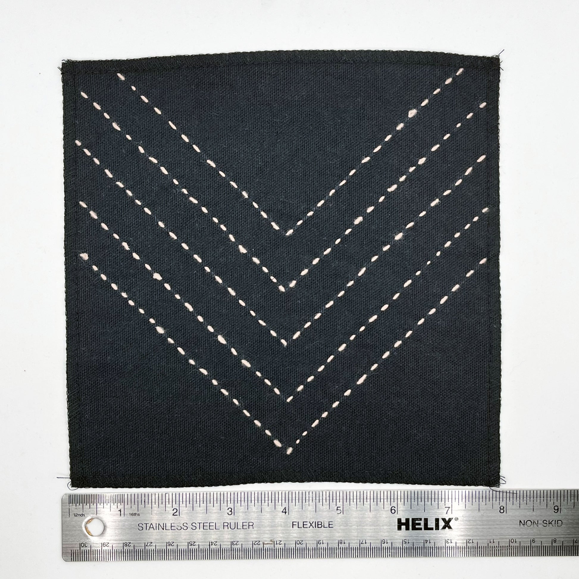 a square patch made out of black canvas, handstitched with rows of peach running stitches in a chevron pattern, with overlocked edges, next to a metal ruler to show a width of 8 inches, on a white background