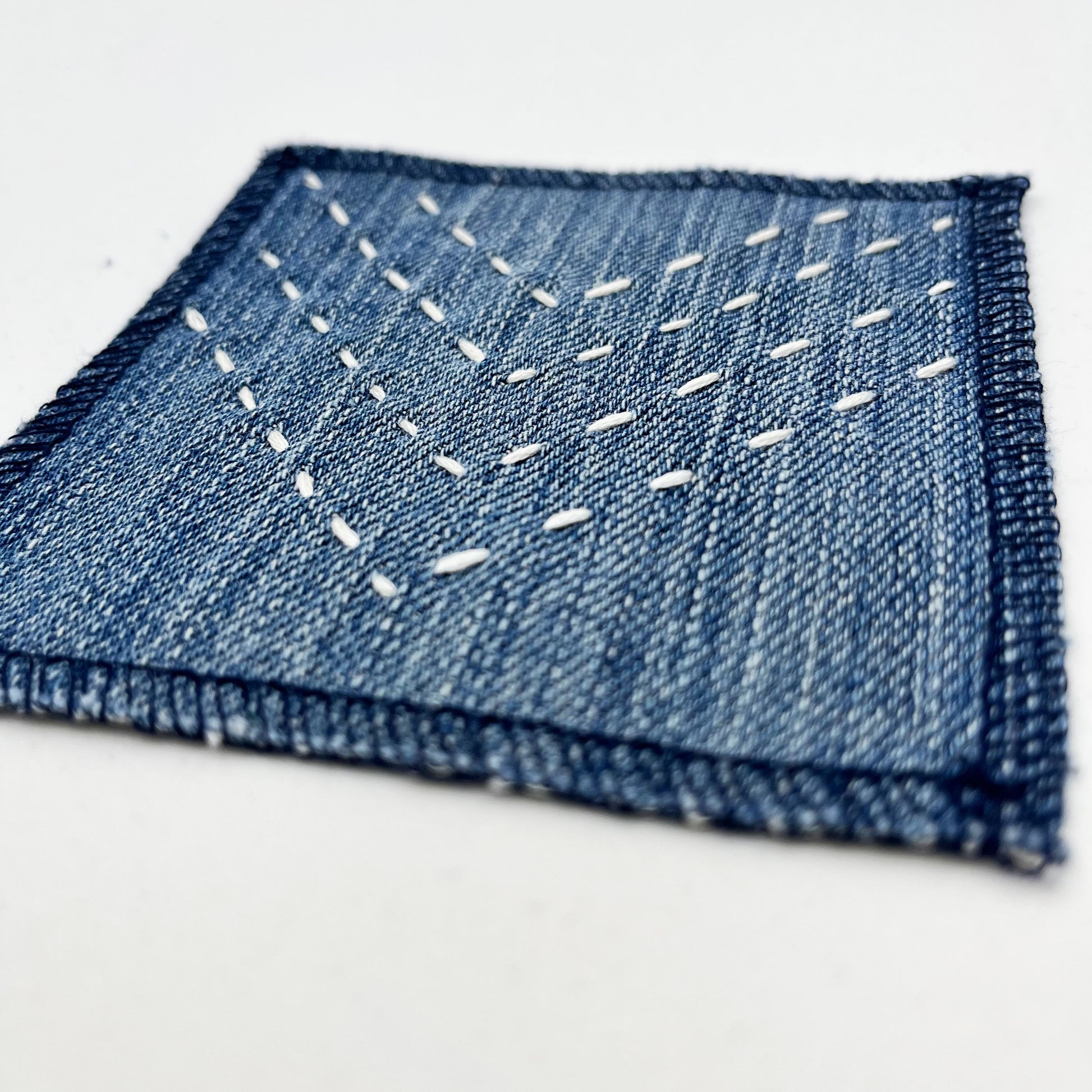 a close up angled view of a square patch made out of denim, handstitched with rows of ivory running stitches in a chevron pattern, with overlocked edges, on a white background