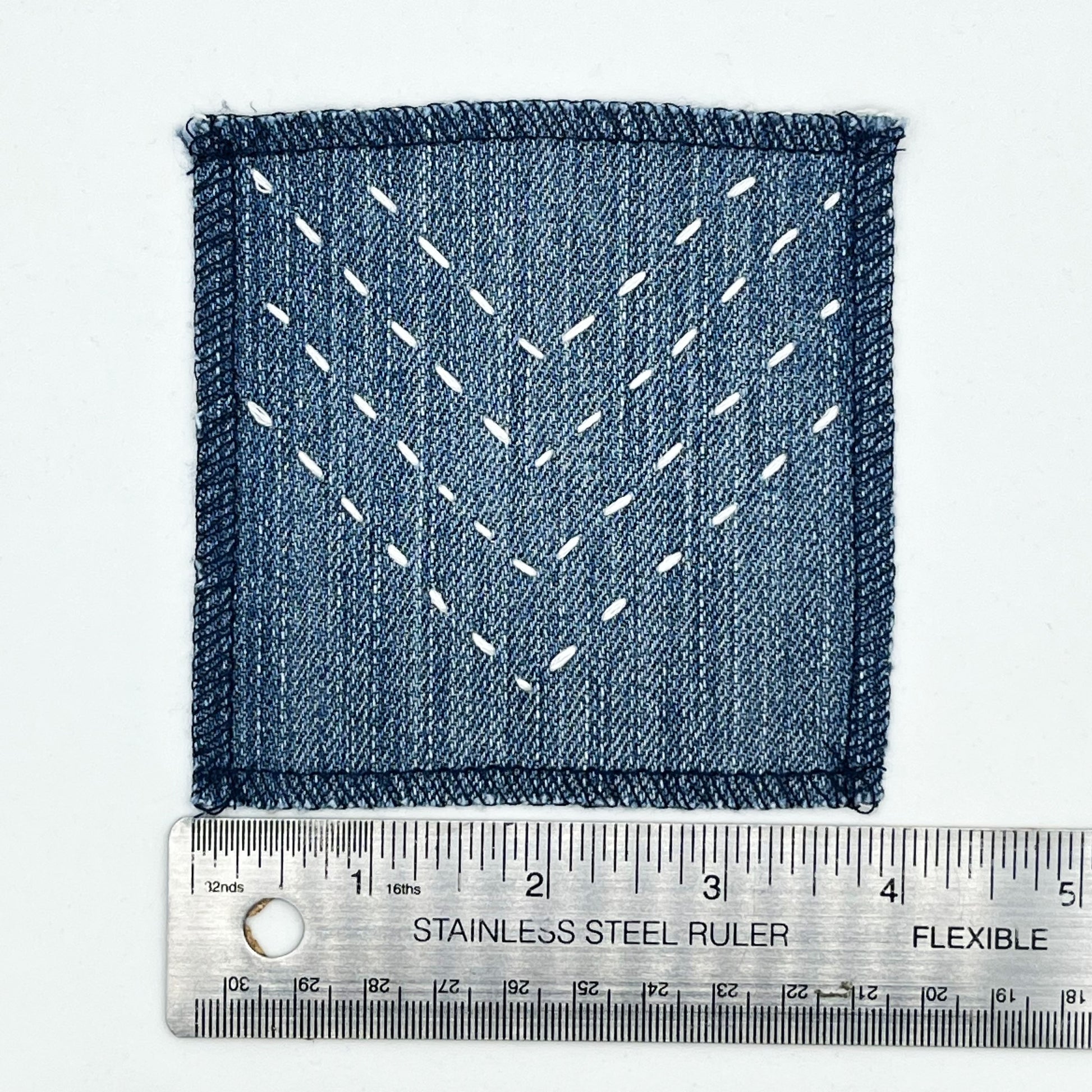 a square patch made out of denim, handstitched with rows of ivory running stitches in a chevron pattern, with overlocked edges, next to a metal ruler showing a width of 4 inches, on a white background