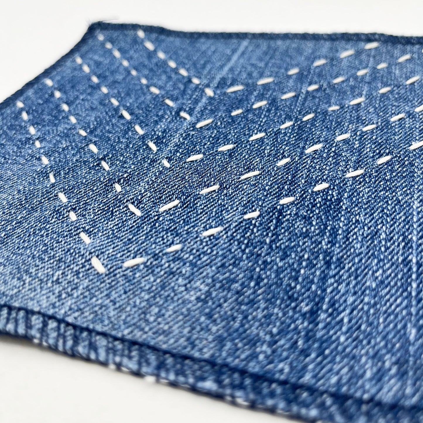 a close up angled view of a square patch made out of denim, handstitched with rows of ivory running stitches in a chevron pattern, with overlocked edges, on a white background