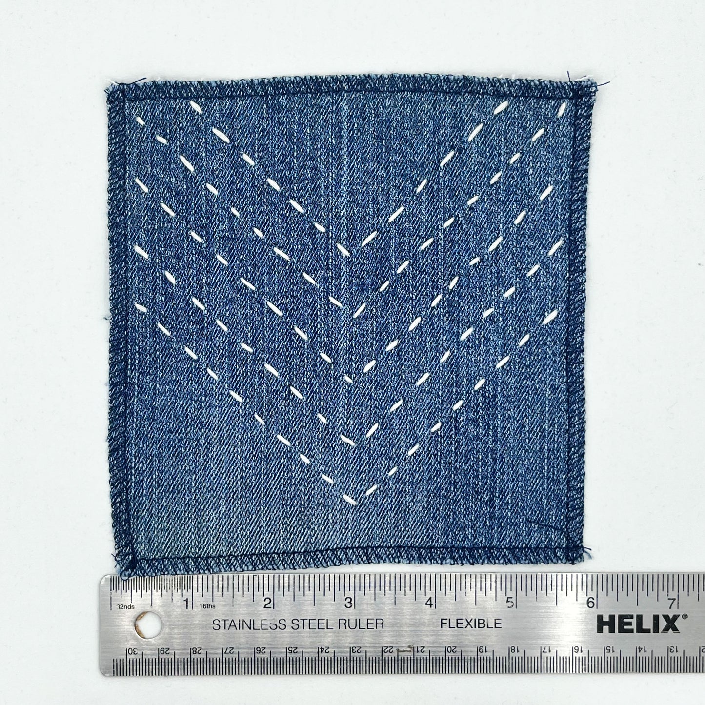 a square patch made out of denim, handstitched with rows of ivory running stitches in a chevron pattern, with overlocked edges, next to a metal ruler showing a width of 6 inches, on a white background