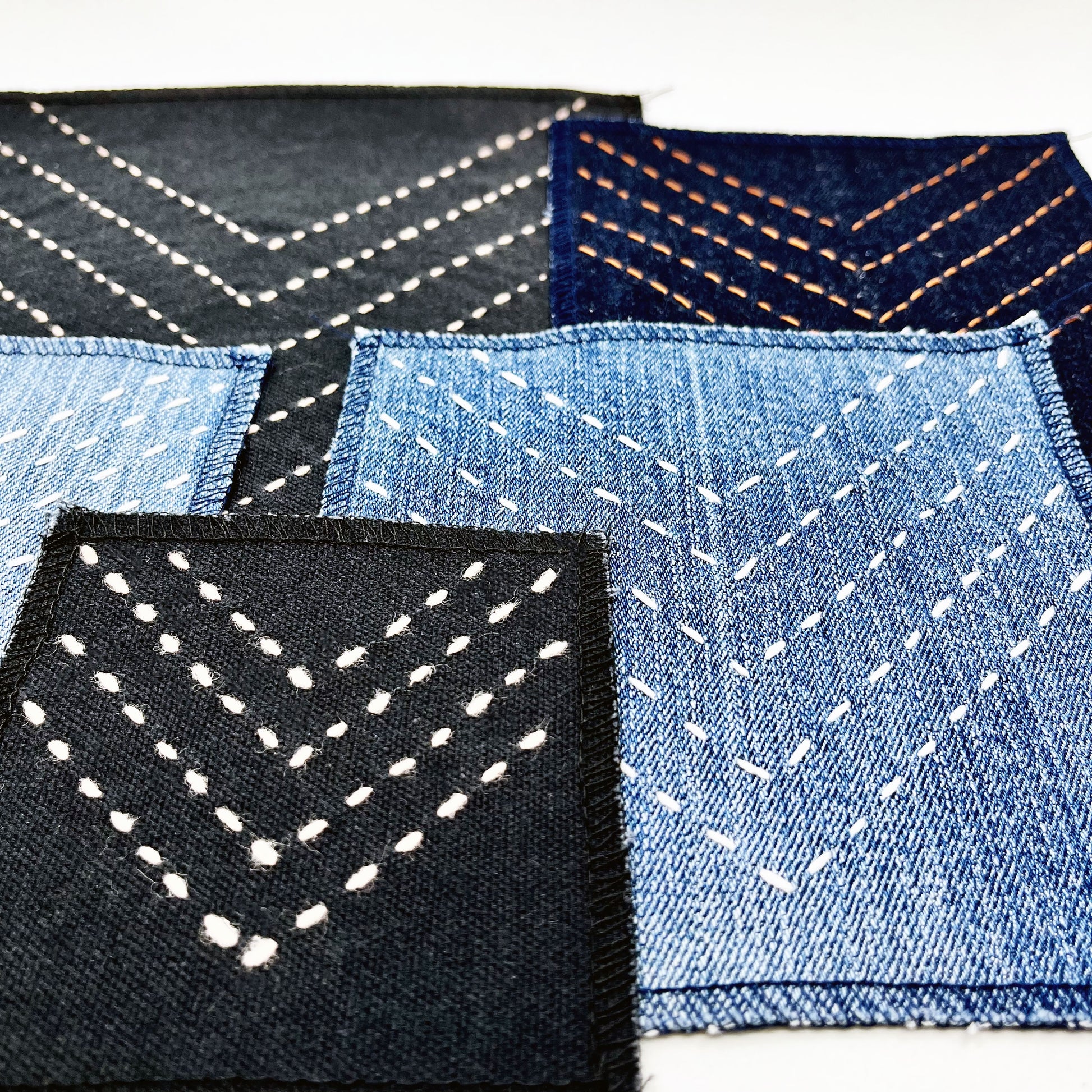a group of square patches made out of denim and black canvas, handstitched with rows of copper (on the dark denim) ivory (on the denim) and peach (on the black) running stitches in a chevron pattern, with overlocked edges, on a white background